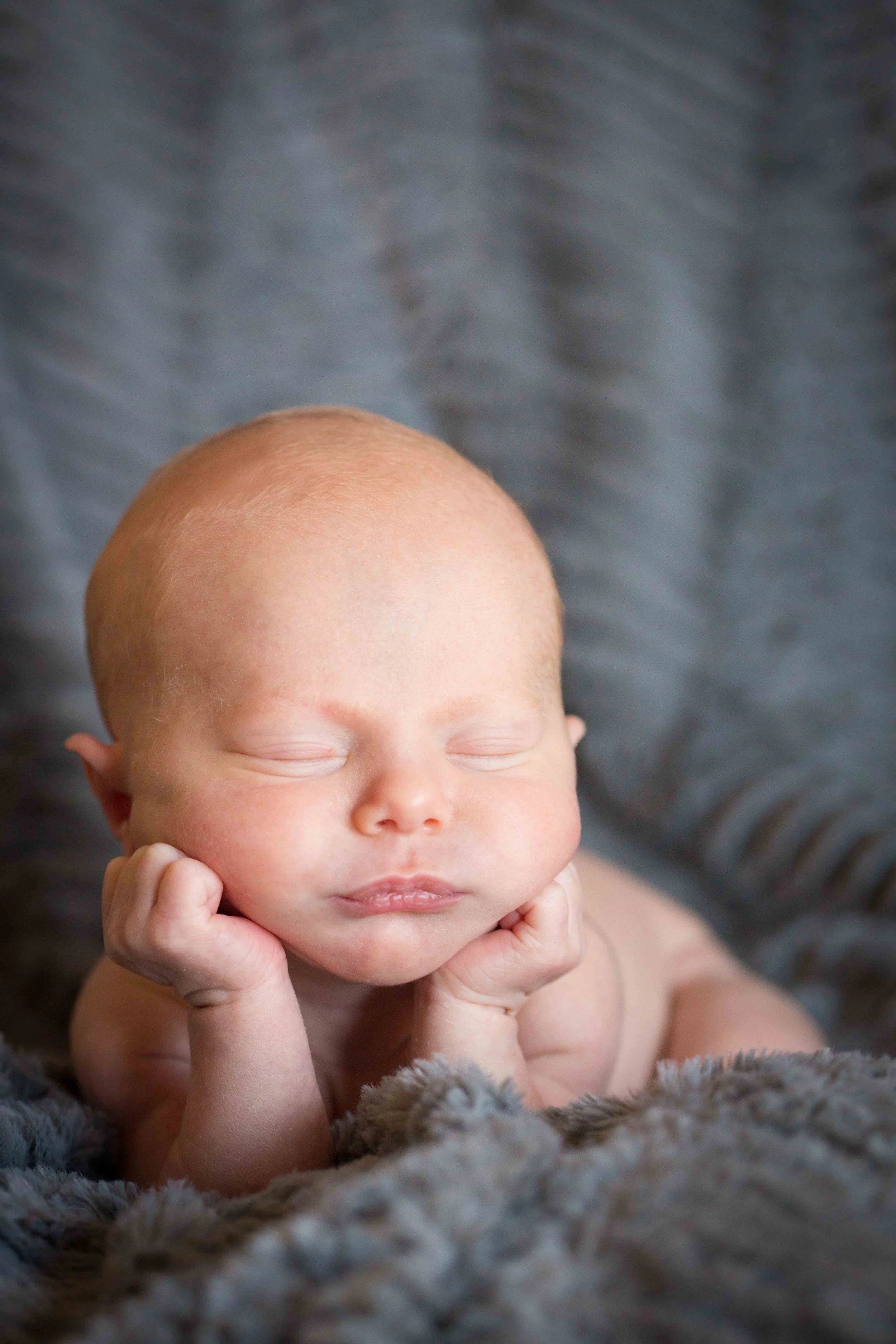 A newborn baby with closed eyes, resting its head on its hands, lying on a soft, gray blanket.