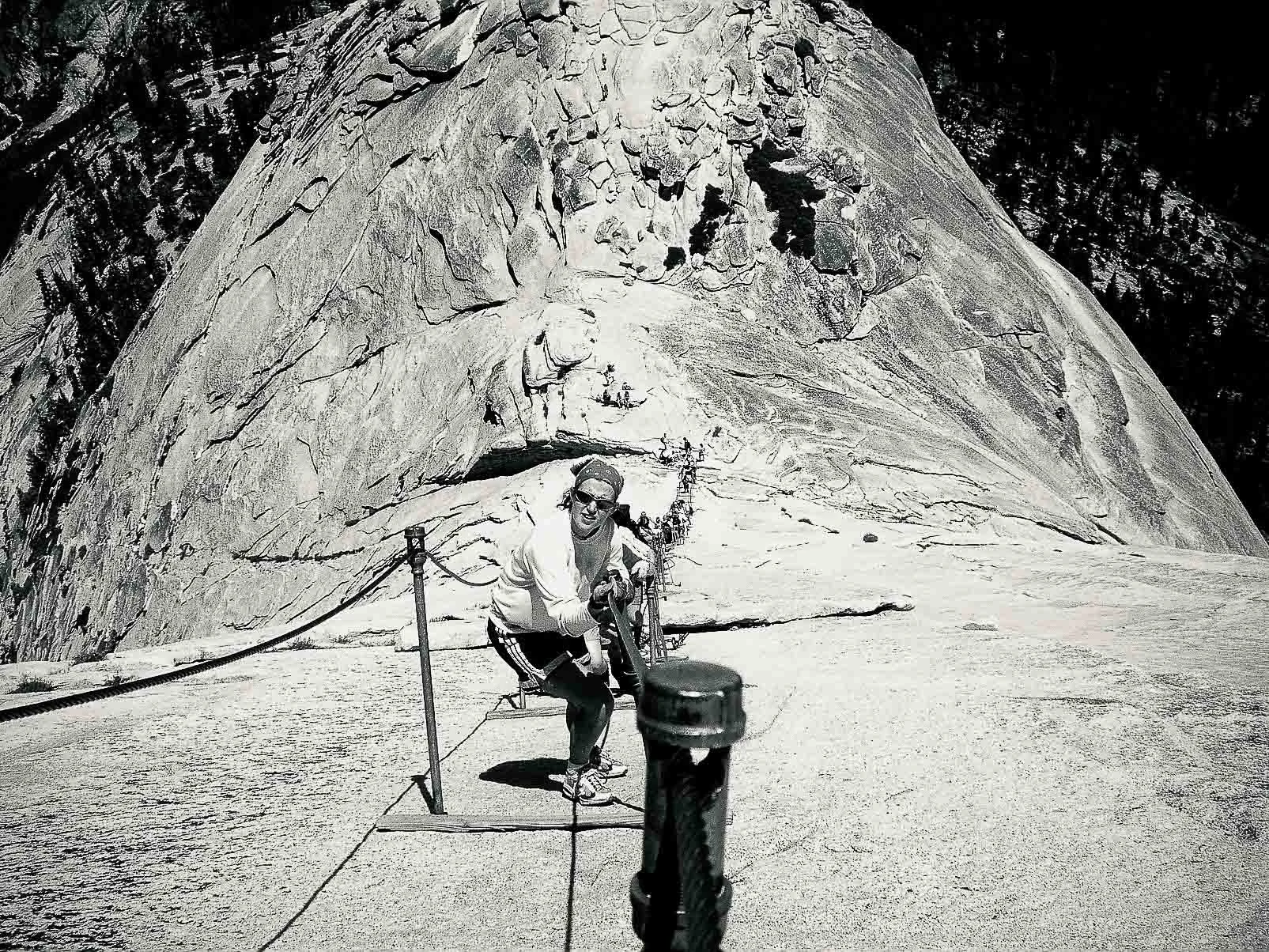 A person in sunglasses and casual clothes holding onto a railing on a mountain trail in front of a large rock formation.