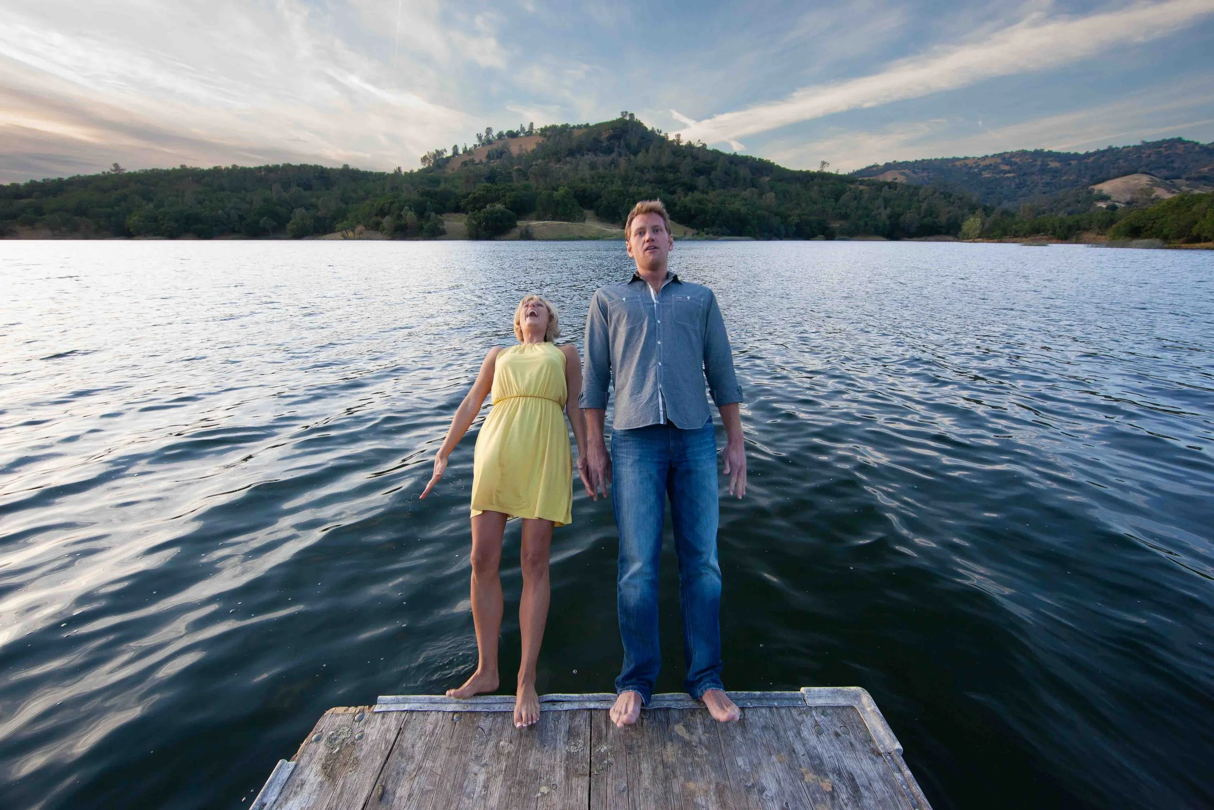 Two people, a woman in a yellow dress and a man in a blue shirt and jeans, standing on a wooden dock over a lake with a mountainous landscape in the background, both looking upwards.