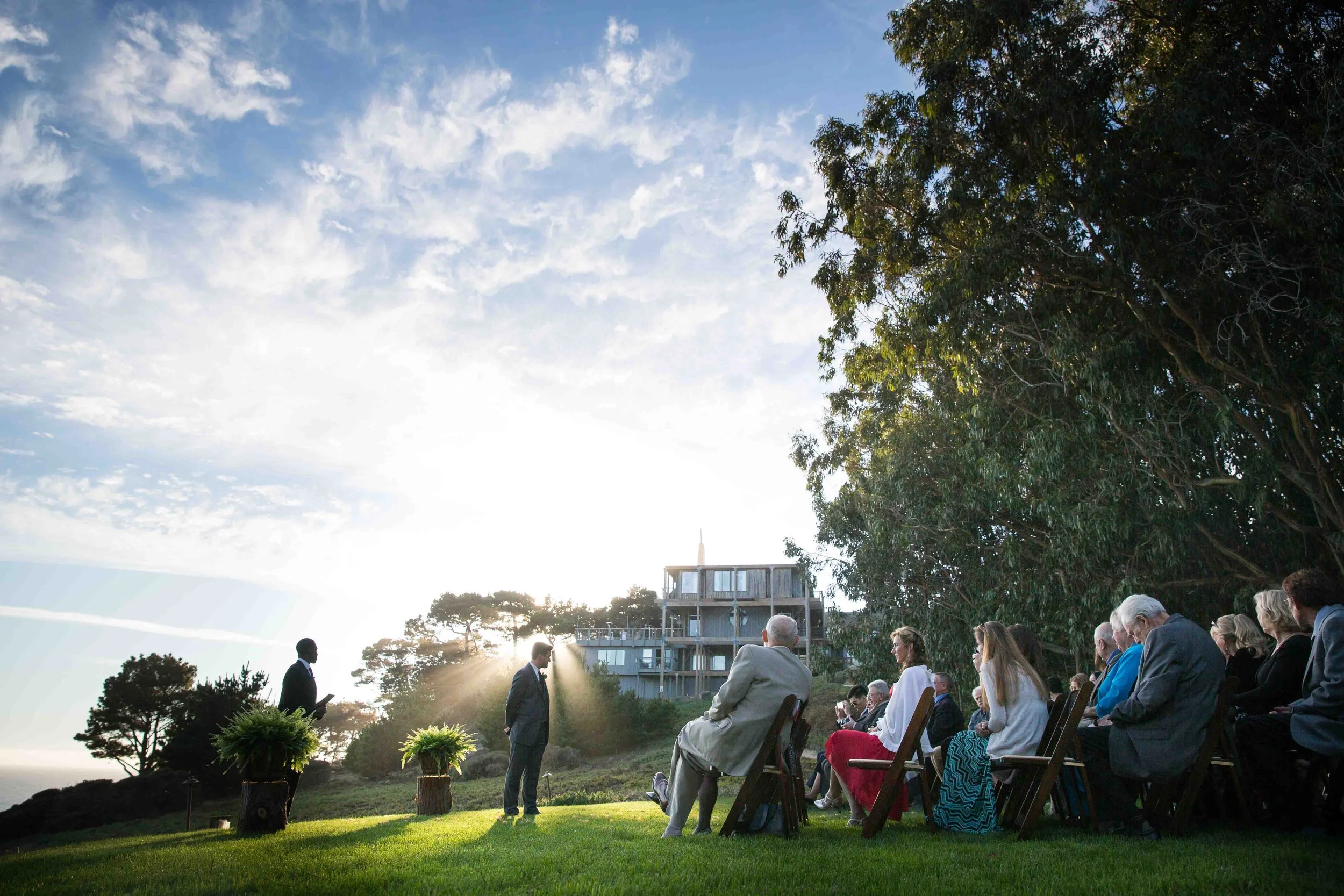 A wedding ceremony outdoors with guests seated on chairs on a grassy area, facing two grooms standing in front of a building at sunset, with sunrays and a partly cloudy sky.