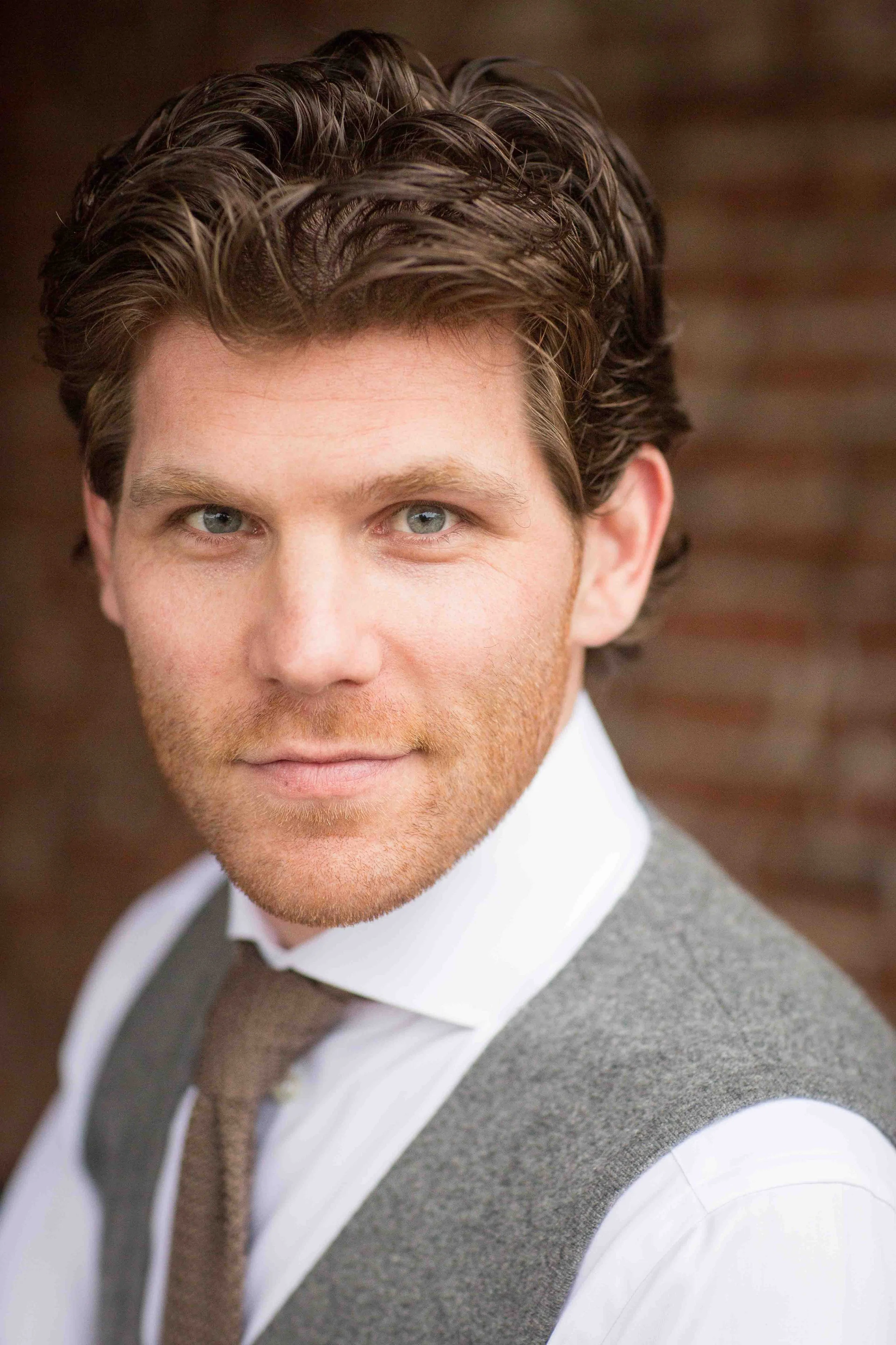 Close-up portrait of a man with wavy brown hair and blue eyes, wearing a white shirt, gray vest, and brown tie.