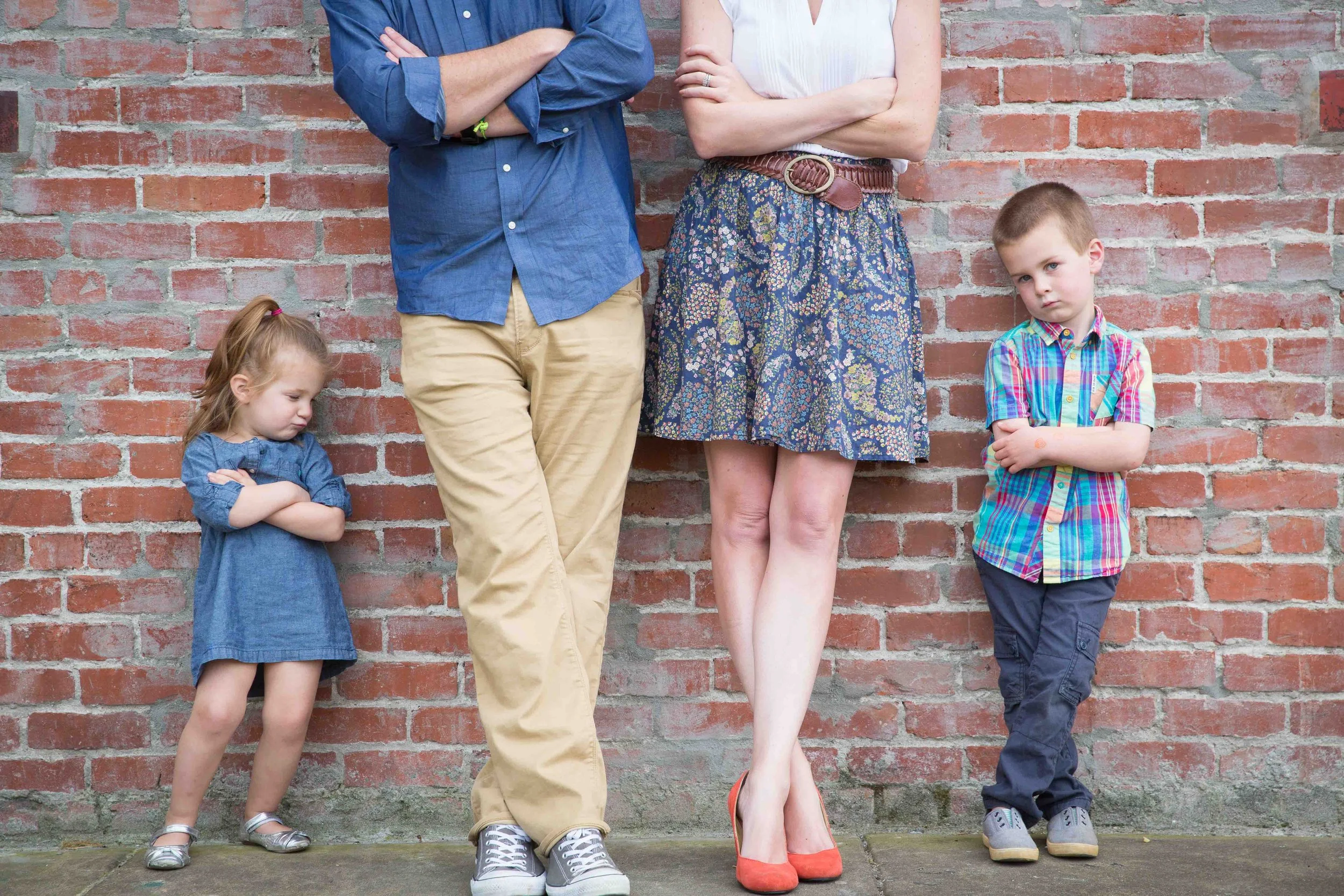 A man, woman, girl, and boy standing against a brick wall with crossed arms, with the girl and boy looking away and crossing their arms.