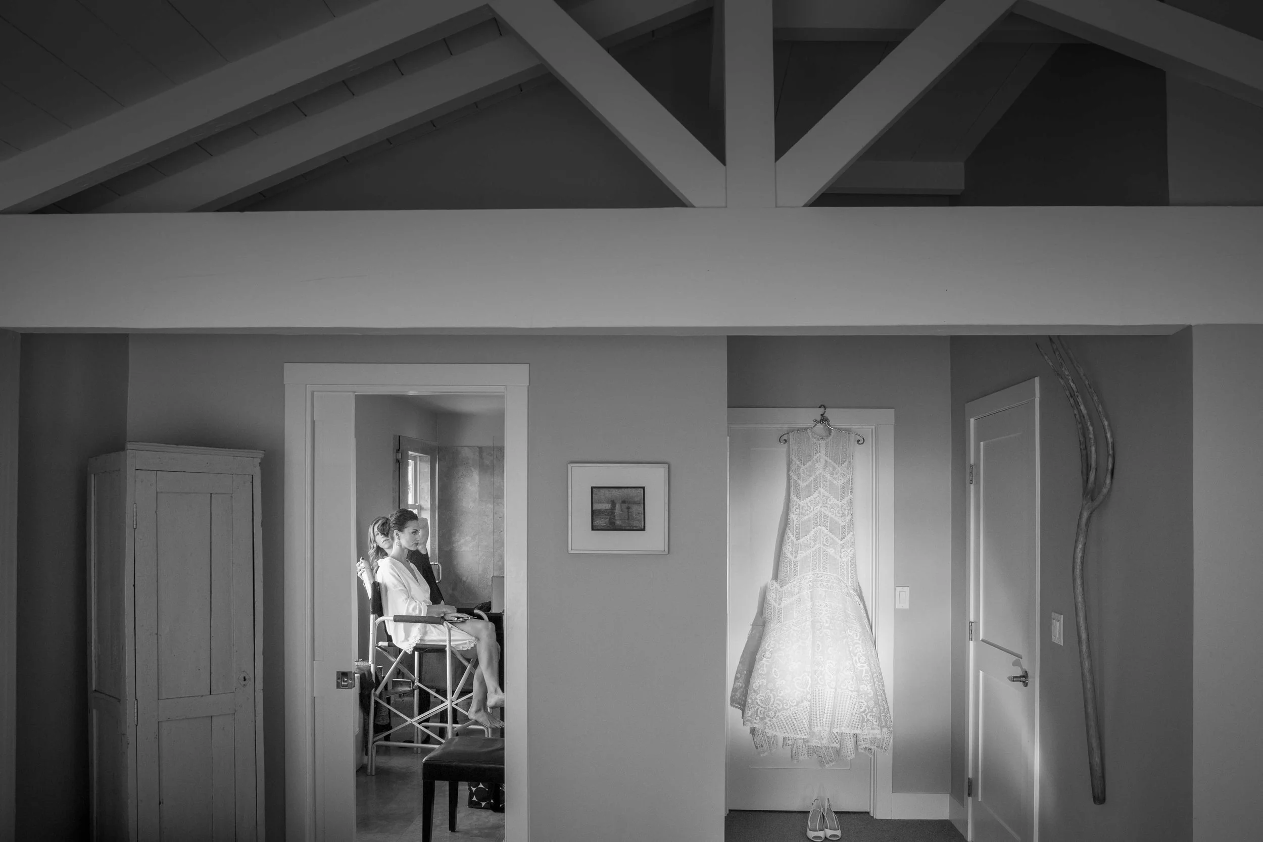 A black and white photo of a woman sitting in a wheelchair in a room, with a door and a window behind her. There is a framed picture on the wall, a lace dress hanging on the door, a walking stick on the wall, and a pair of shoes on the floor.