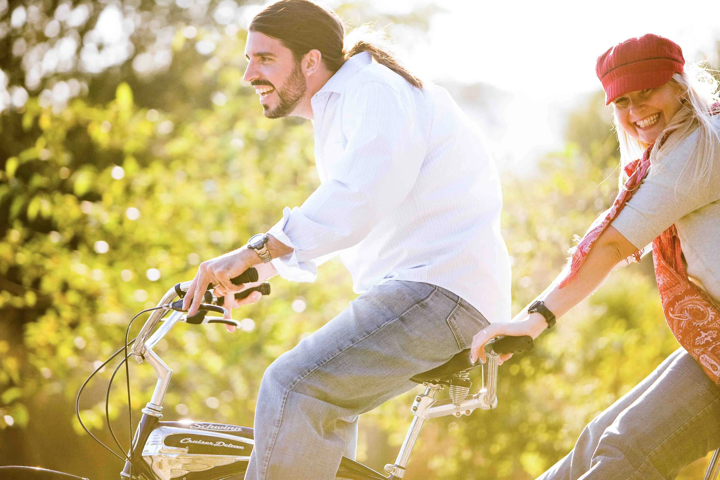 A man and woman riding a tandem bicycle outdoors in a natural setting during daytime, both smiling and enjoying the ride.