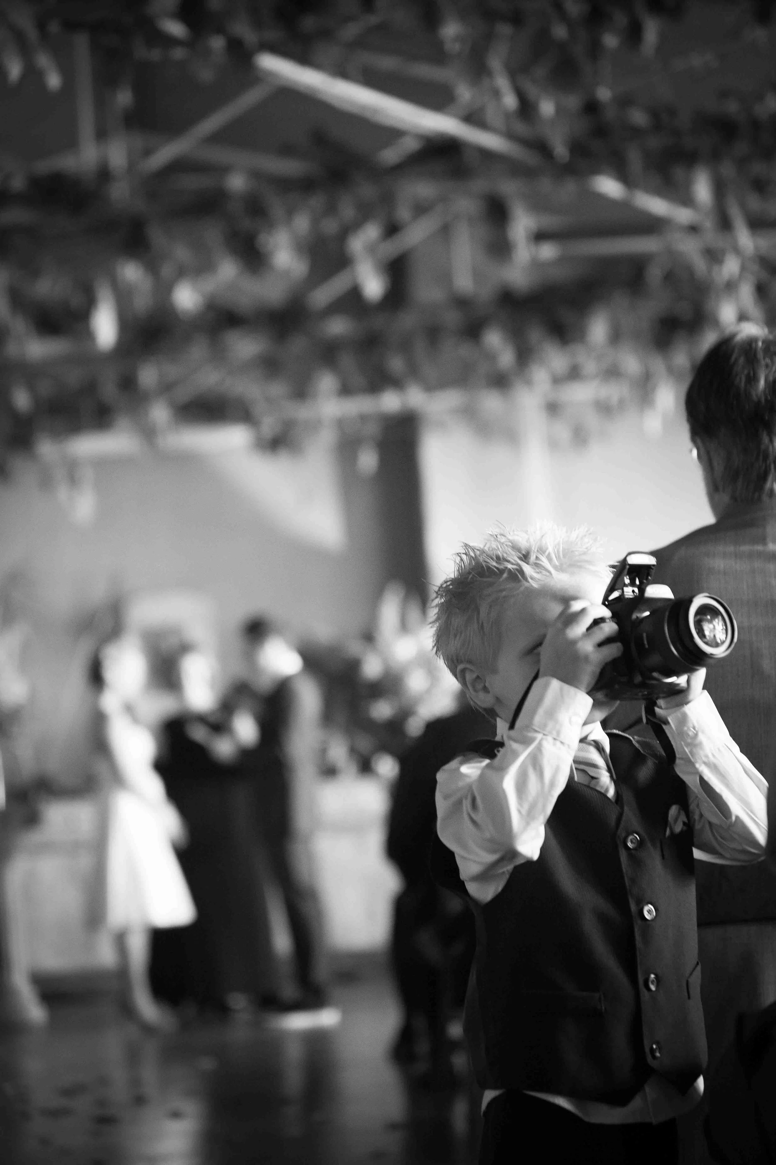 A young boy with spiky blonde hair in a vest and dress shirt holding a camera, taking a photo at a formal event, with blurred wedding guests in the background.