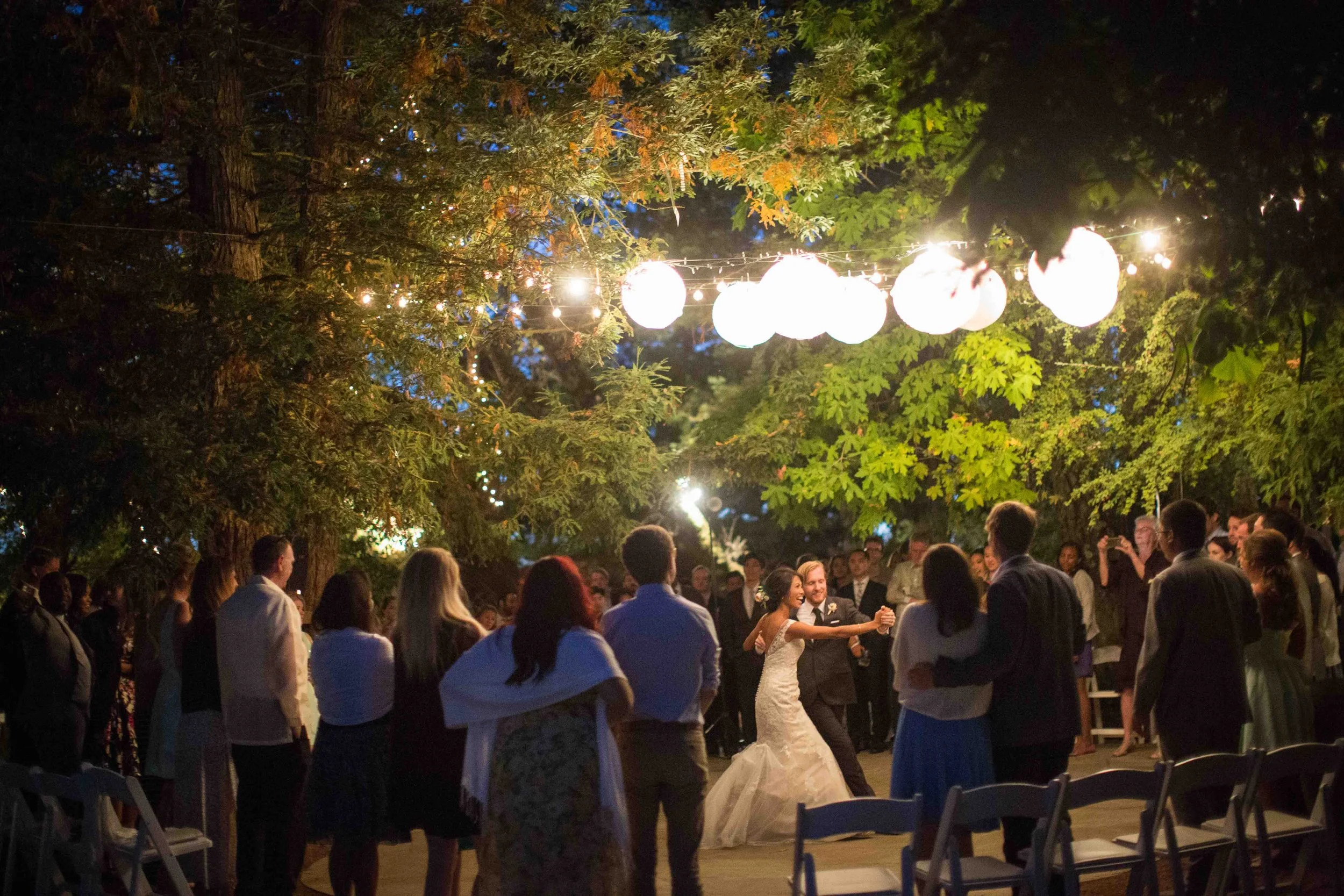 A group of people dancing at an outdoor wedding reception under hanging paper lanterns and string lights, surrounded by trees at night.