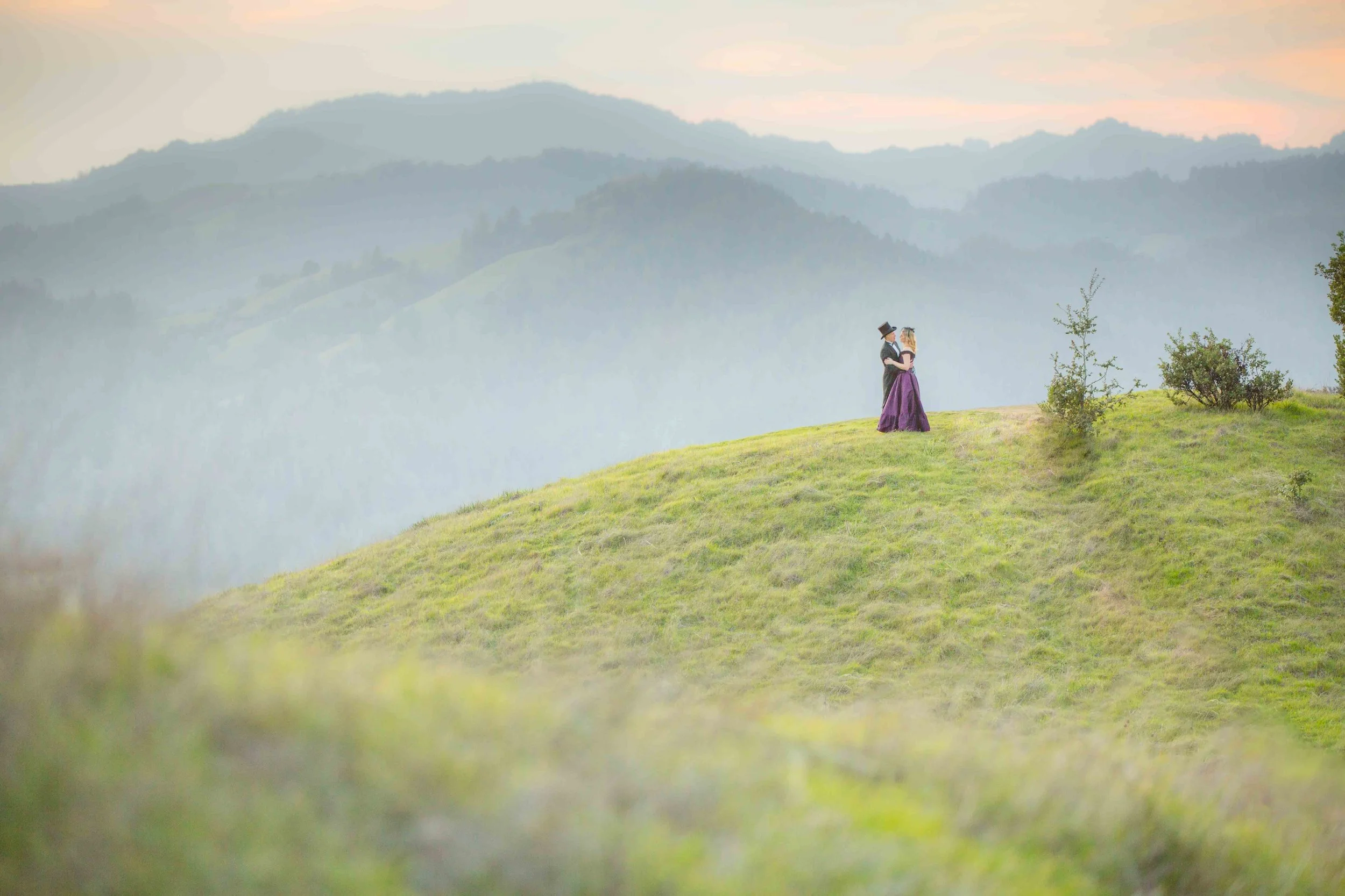 A couple dressed in vintage clothing standing on a grassy hill embracing each other with a backdrop of misty mountains and soft sky at sunset.