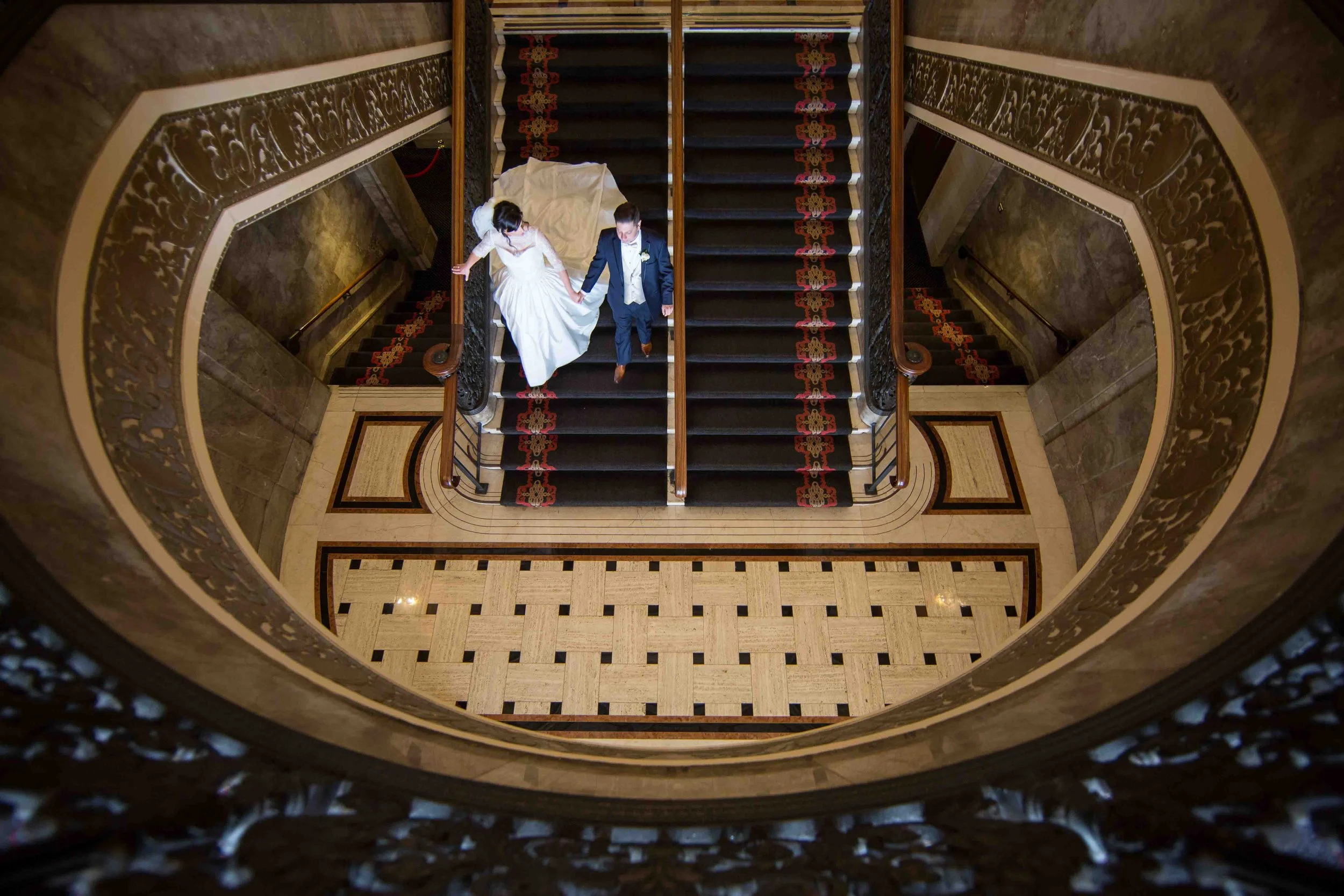 Overhead view of a bride and groom walking hand in hand down a staircase inside a grand building with ornate wall designs and a patterned floor.