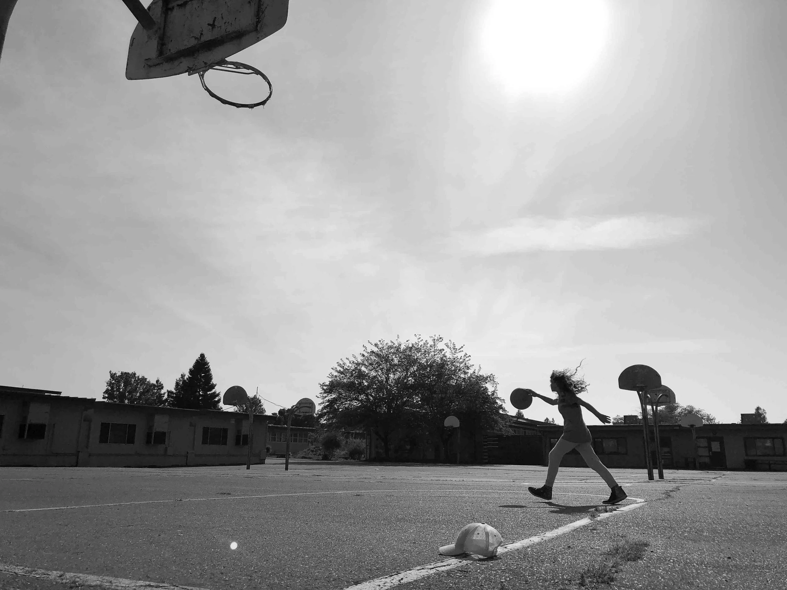 A young girl with curly hair playing basketball on an outdoor basketball court, with a backboard and hoop visible and a baseball cap on the ground. The photo is taken in black and white during daytime, with trees and school buildings in the backgroun