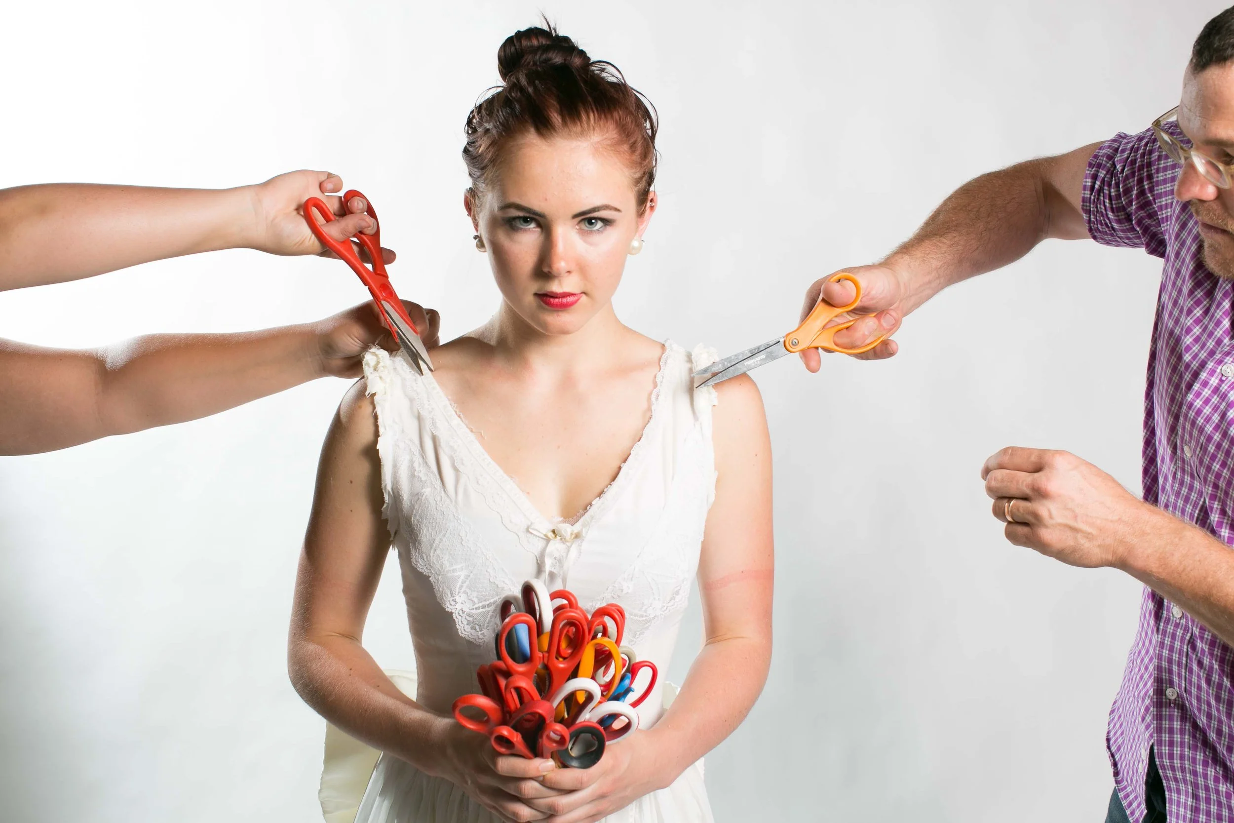 A woman holding scissors with multiple pairs of scissors being used by two men to cut her clothing at her shoulders.