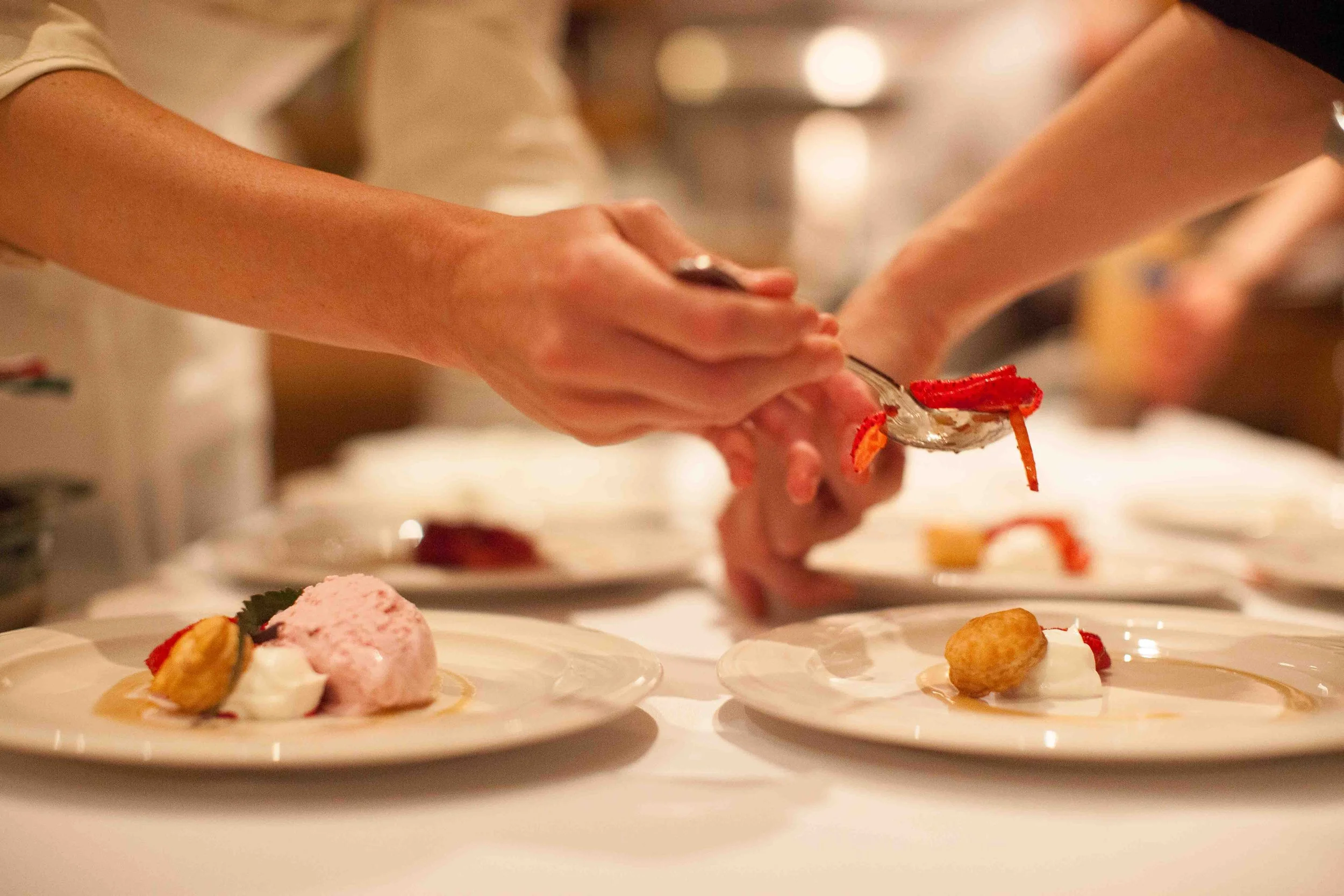 Chef garnishing desserts on plates, with hands using tongs and spoon, in a professional kitchen setting.
