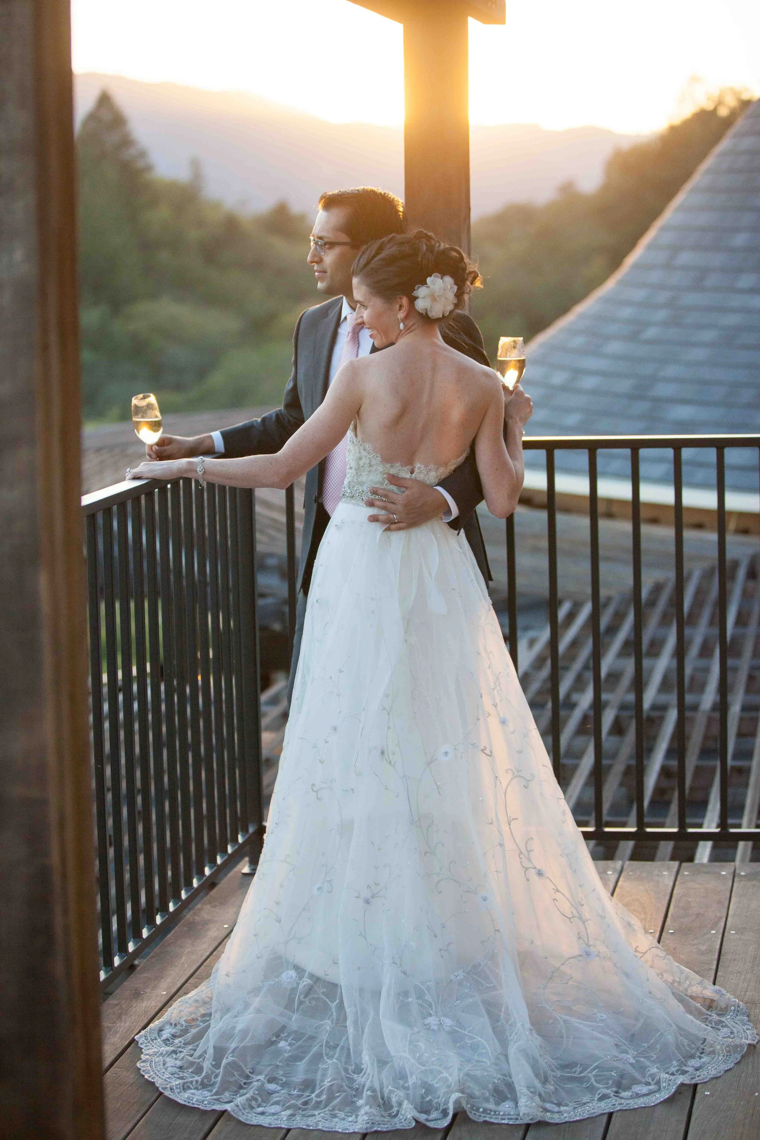 A newlywed couple sharing a romantic moment on a balcony at sunset, with the bride in a white wedding dress and the groom in a dark suit, both holding glasses of champagne.