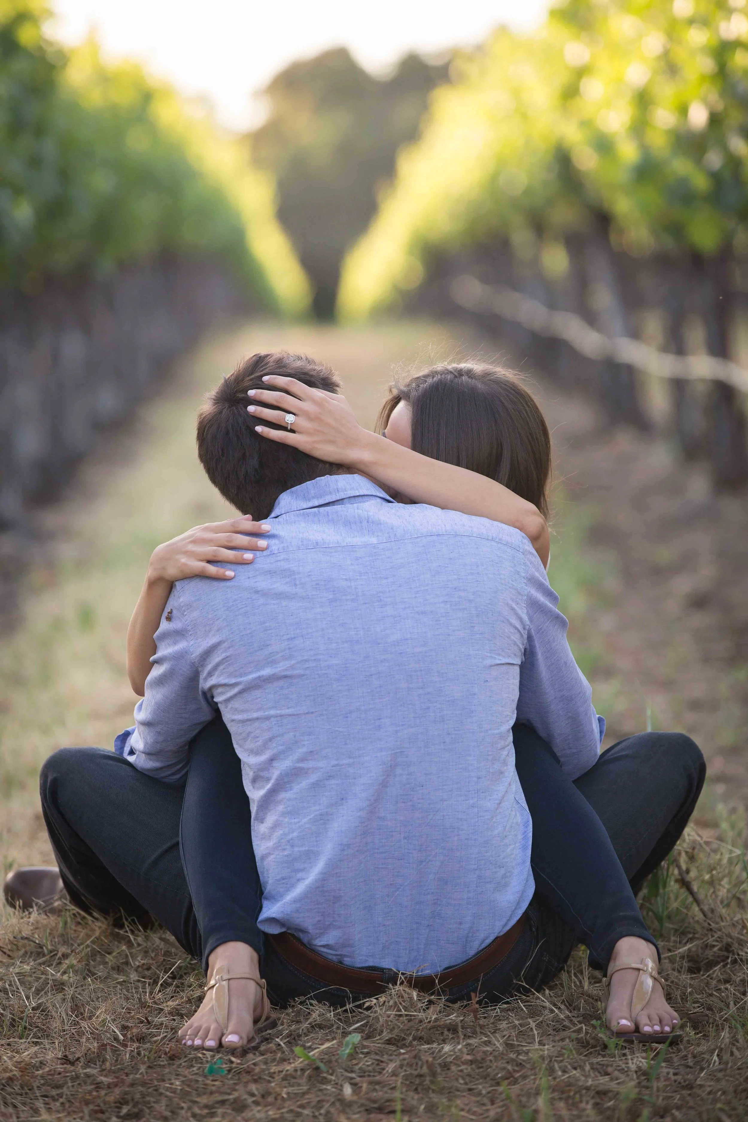 A couple sitting closely on the ground in a vineyard, embracing and kissing with trees and sunlight in the background.