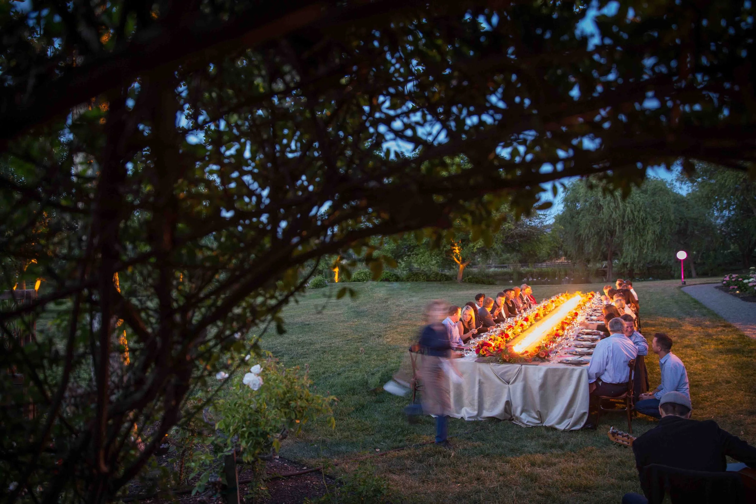 A long outdoor dinner table with people dining, illuminated by a glowing light, set in a garden during dusk, with trees and pathway in the background.