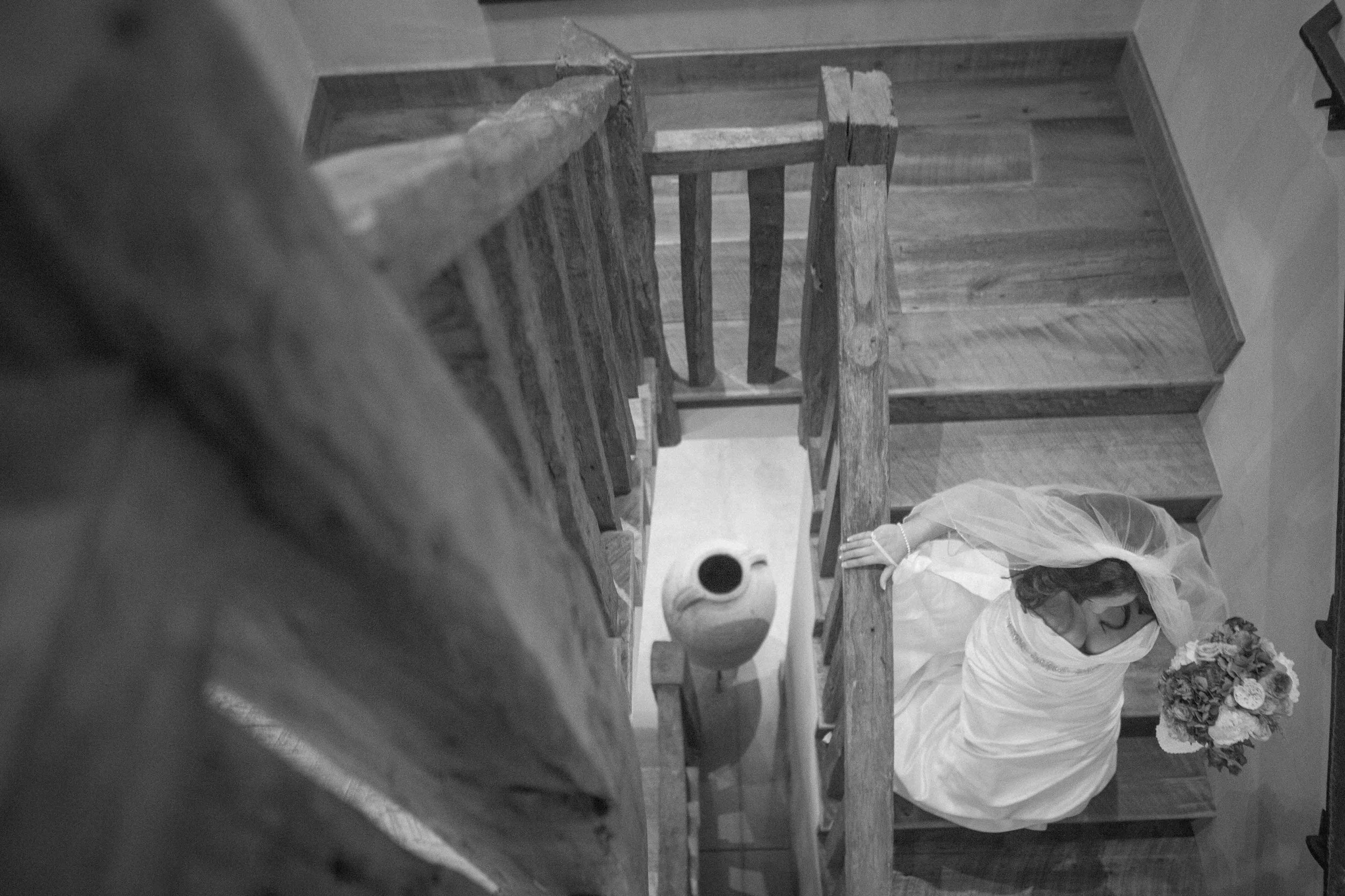 Black and white photo taken from the top of a staircase showing a woman in a wedding dress holding a bouquet, looking up, with the camera angle focusing on her from above.