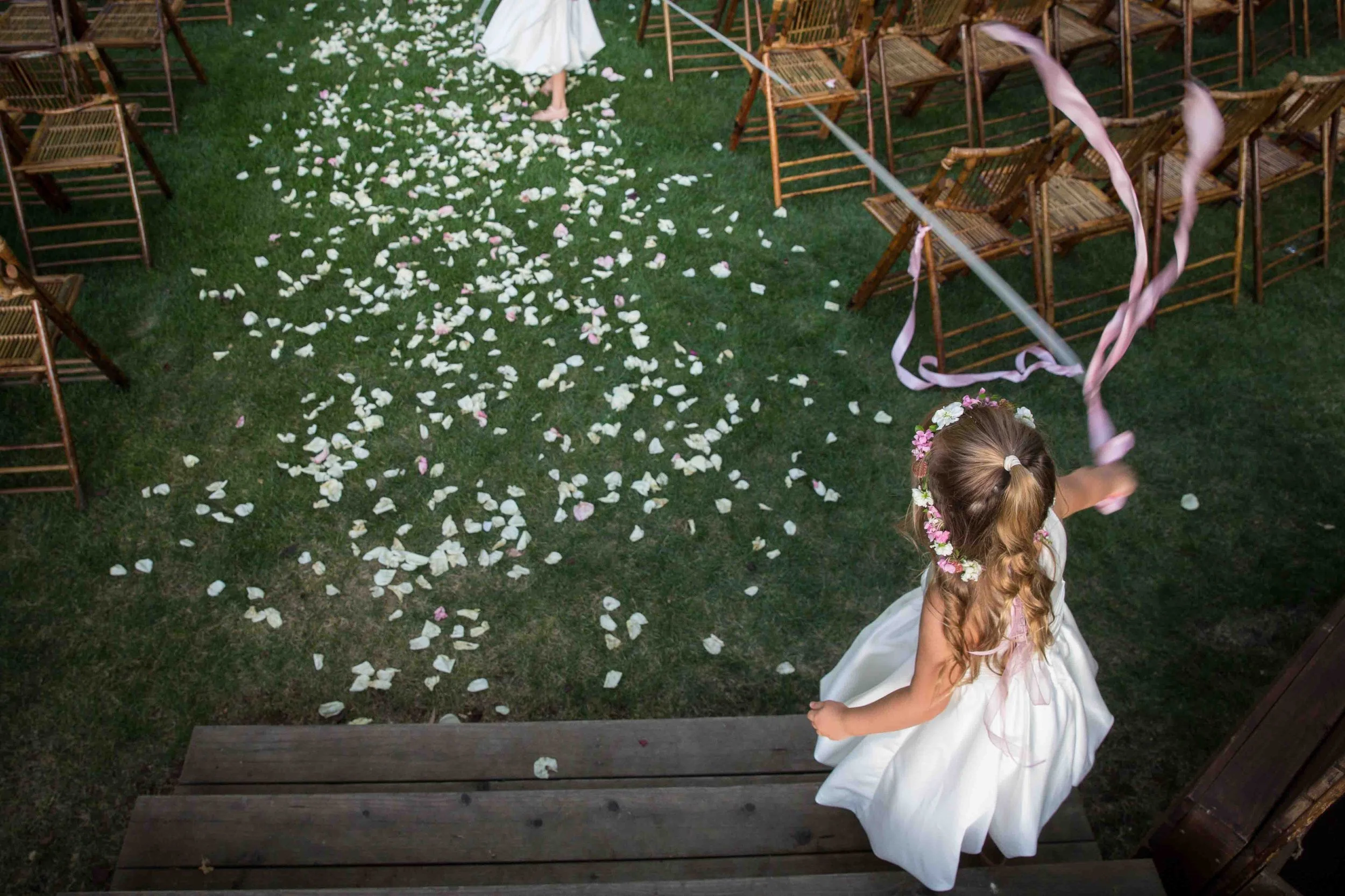 A young girl in a white dress and floral headband holding pink ribbons, celebrating at a wedding, with flower petals scattered on the grass and wooden chairs arranged for guests.