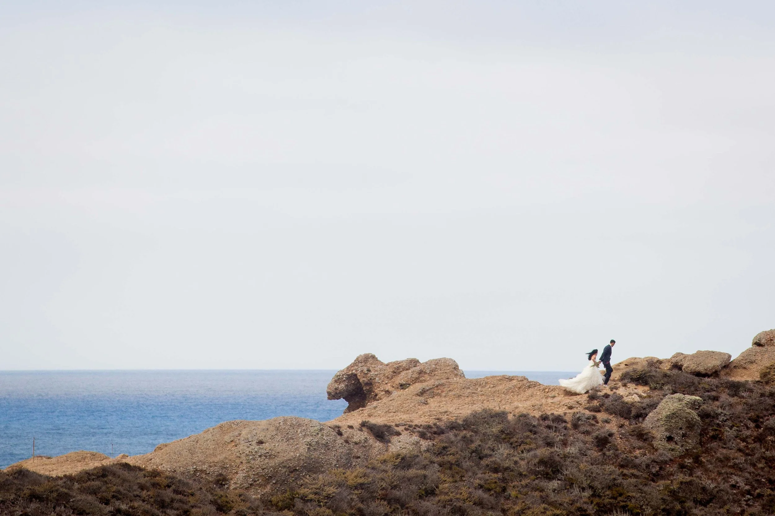 A bride and groom in wedding attire walking on rocky coastal terrain near the ocean under an overcast sky.
