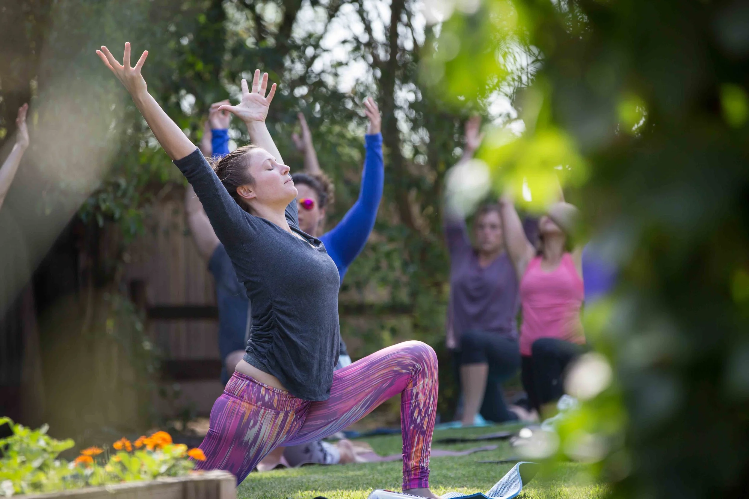 A group of women practicing yoga outdoors in a lush garden, with one woman in the foreground performing a yoga pose with arms raised and eyes closed.
