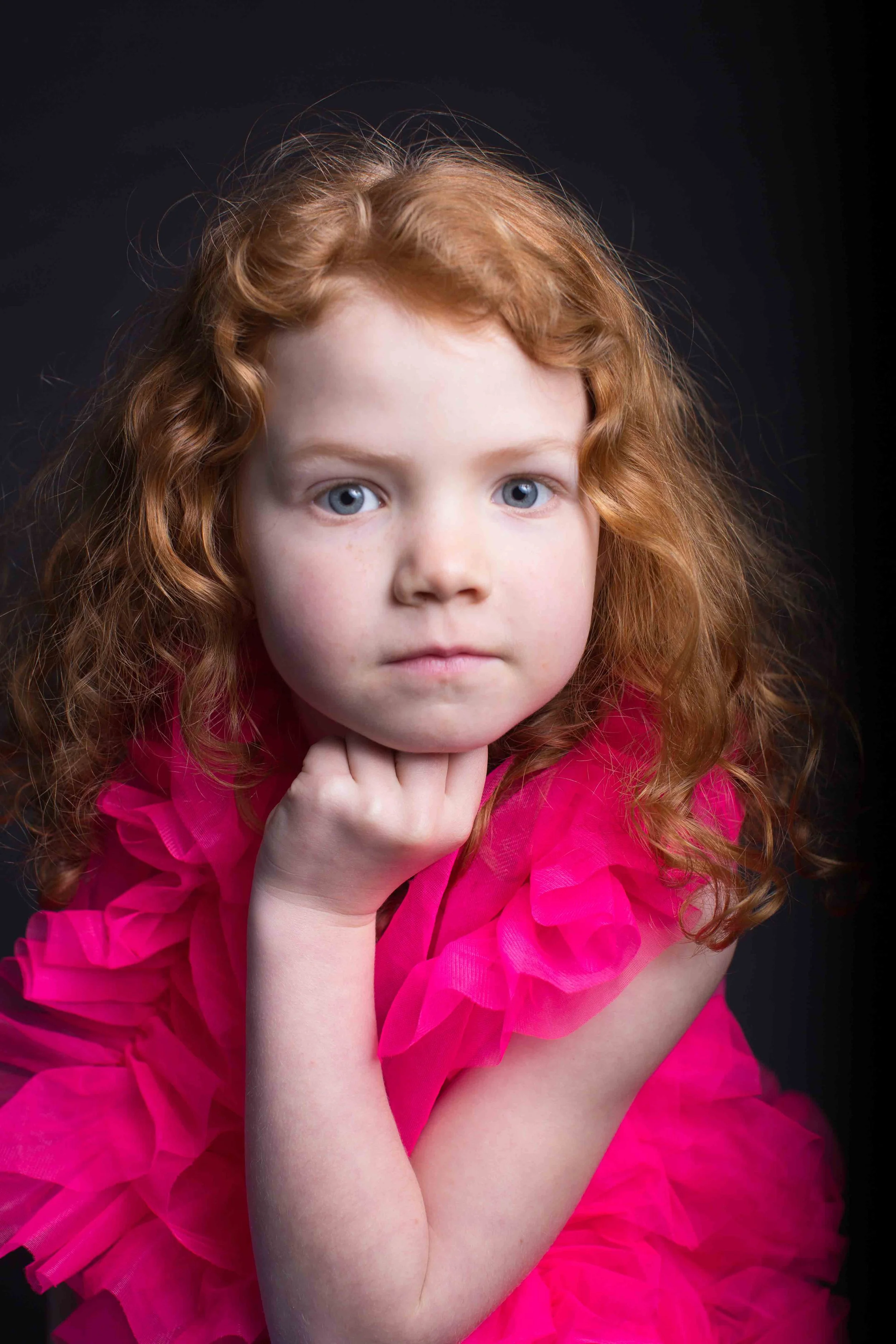 Portrait of a young girl with curly red hair and blue eyes, wearing a bright pink ruffled dress, resting her chin on her hand against a dark background.