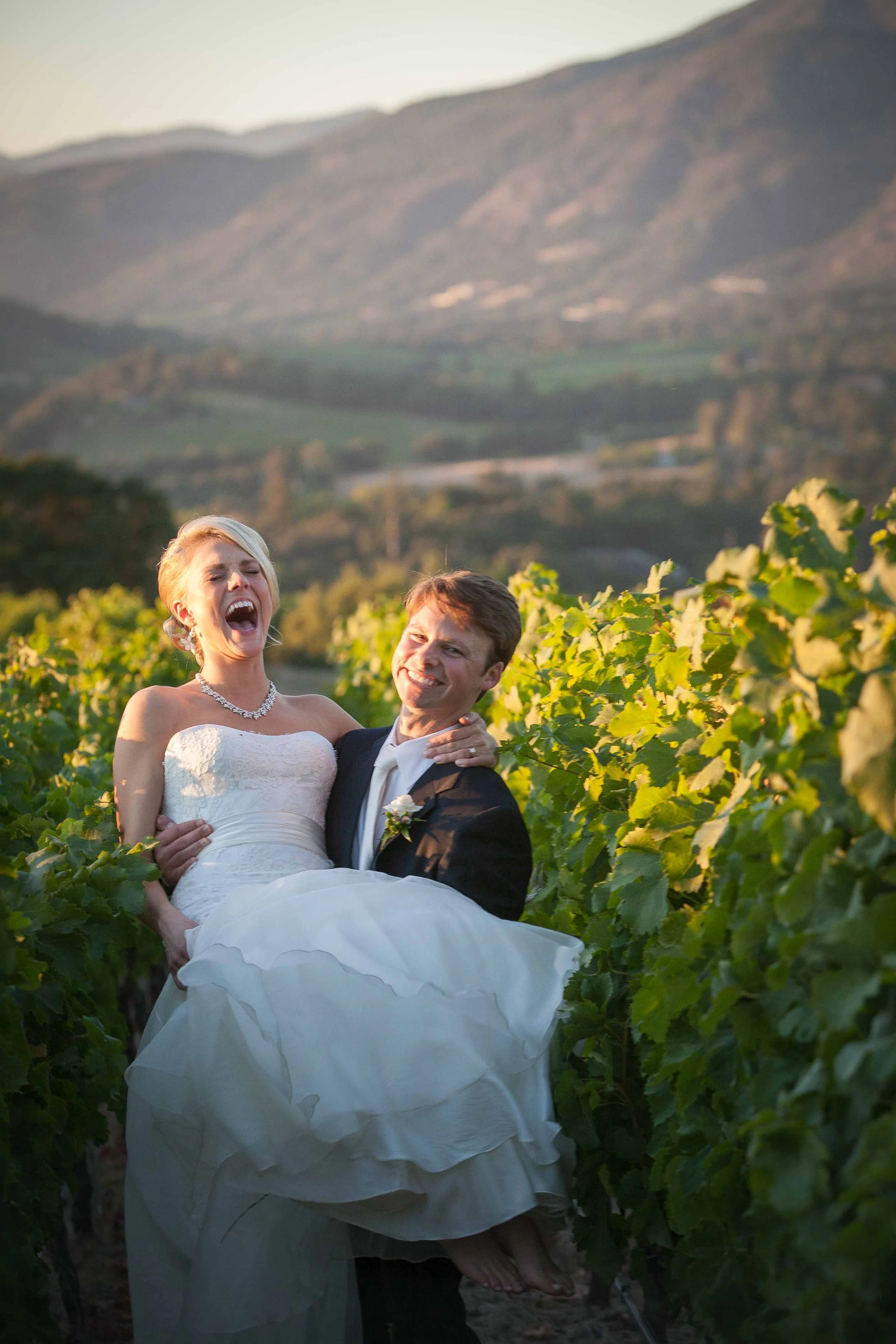 A bride in a white wedding dress and a groom in a black suit are happily posing outdoors among green vineyard rows with a mountain range in the background, during sunset or golden hour.