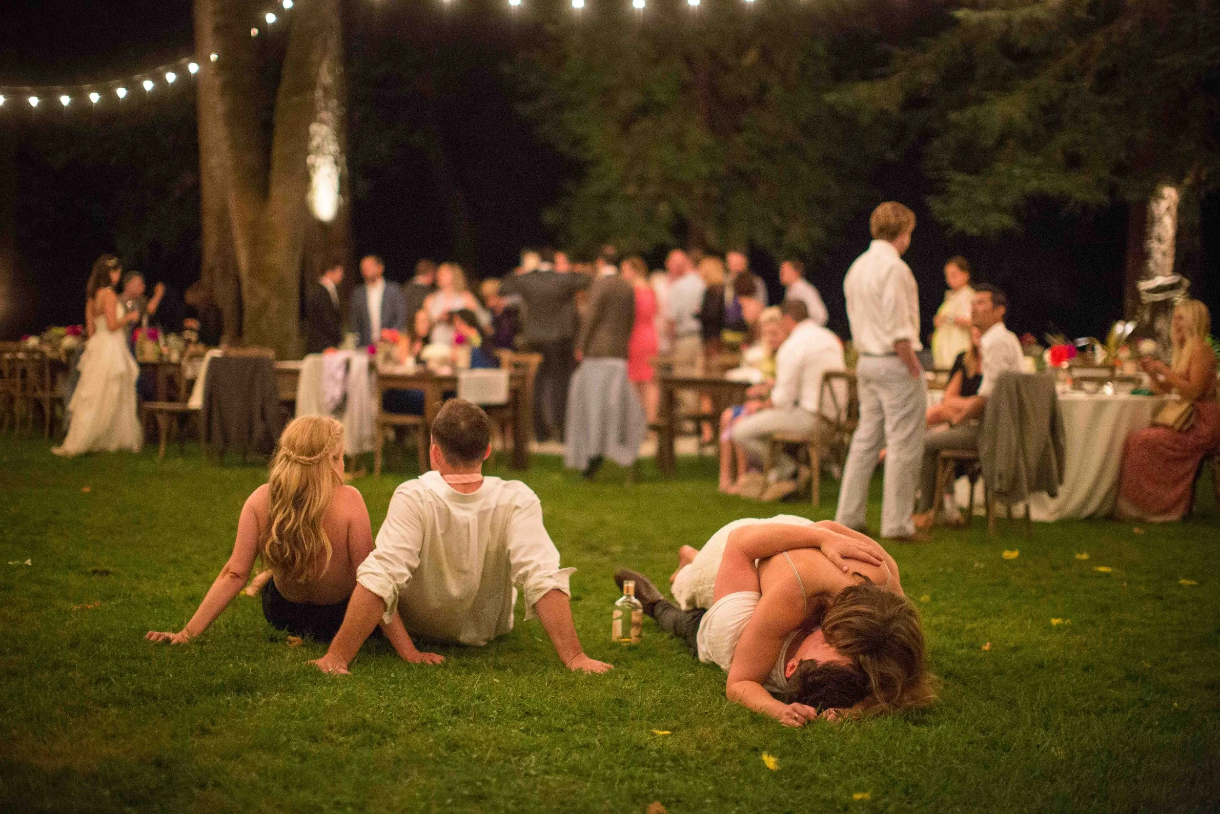 People at a wedding reception outdoors at night, with string lights overhead. Some guests are seated at tables, while others dance and relax on the grass, including a couple kissing and a woman lying on top of a man.