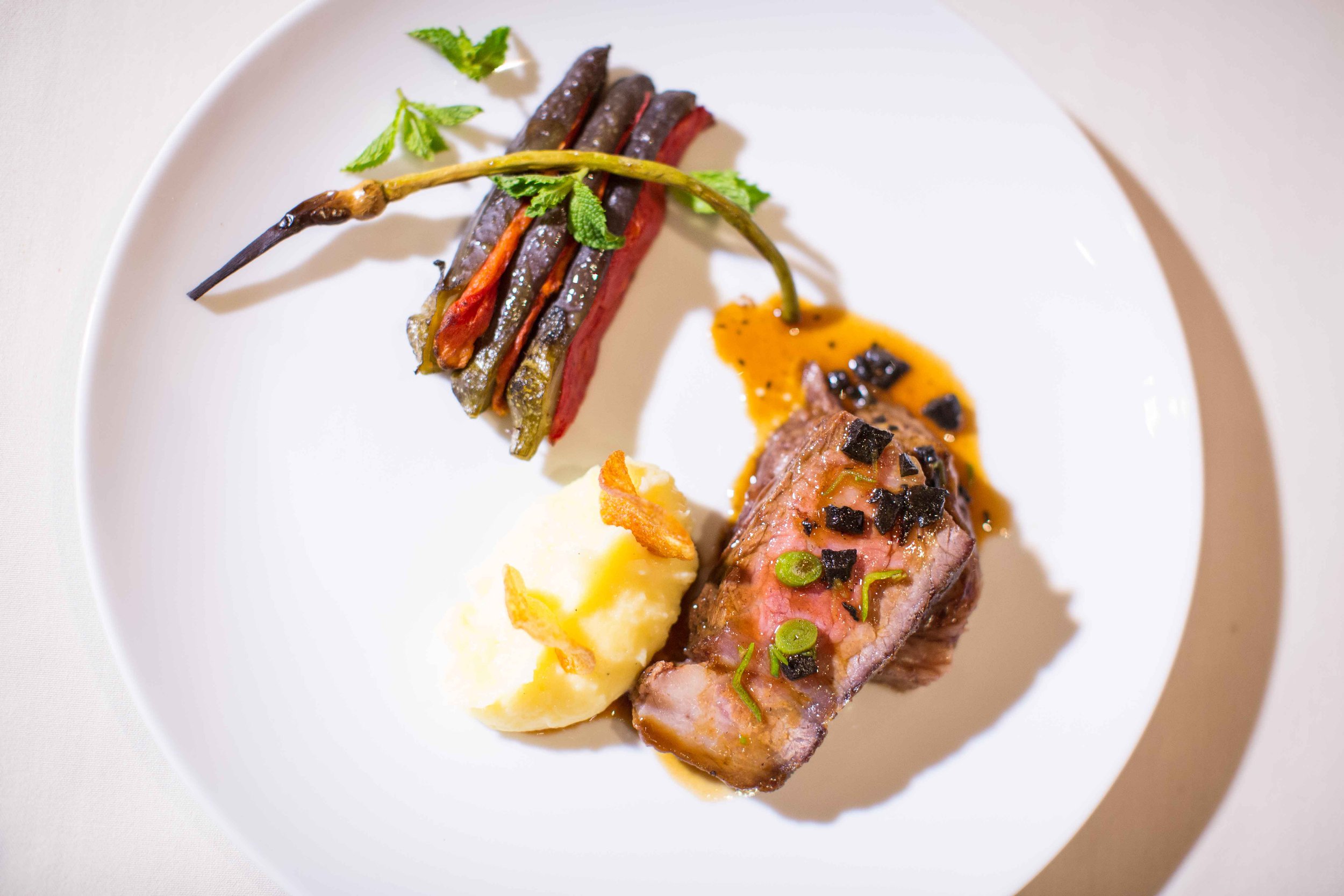 A plated gourmet meal with a slice of cooked steak topped with sauce and green onions, a mound of mashed potatoes with crispy onion bits, and a skewer of colorful grilled vegetables including red, yellow, and green peppers, garnished with a sprig of 