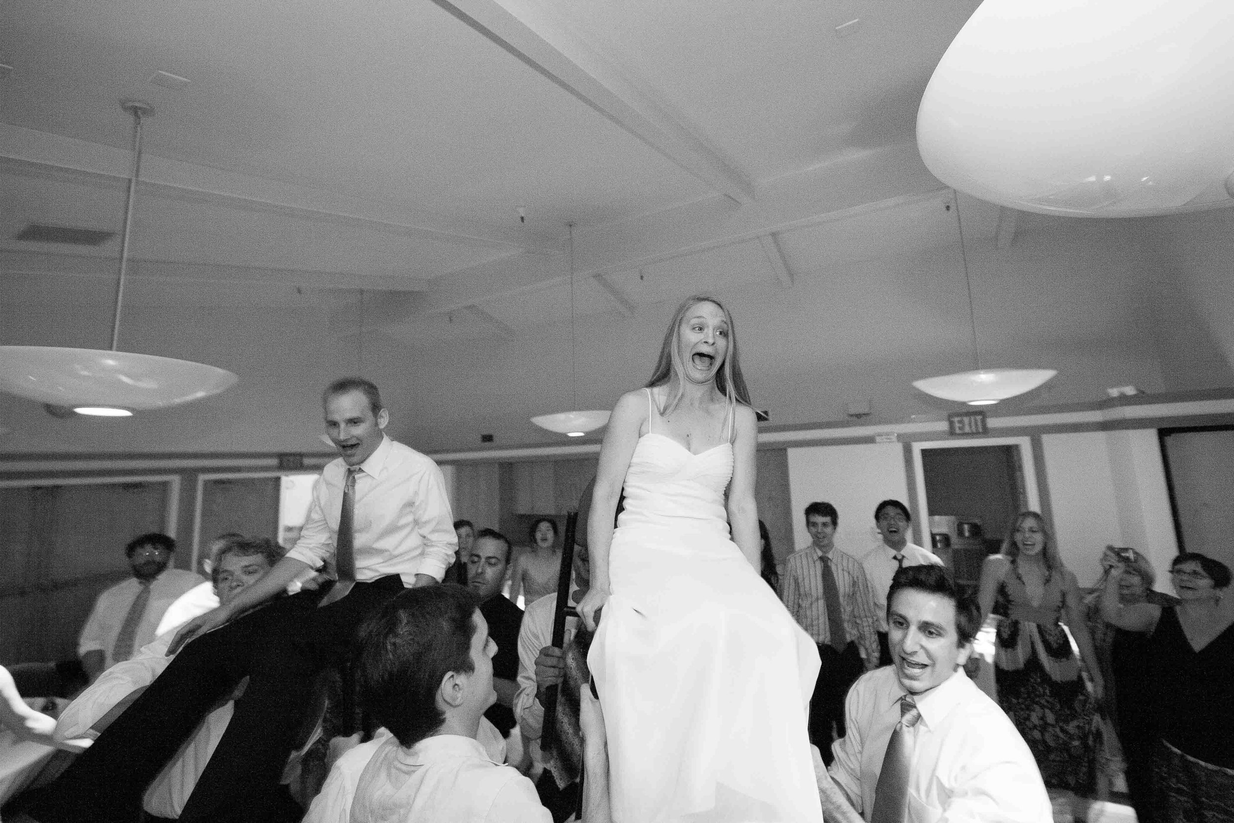 A wedding reception where a woman in a wedding dress is being lifted up on a chair, surrounded by laughing guests.