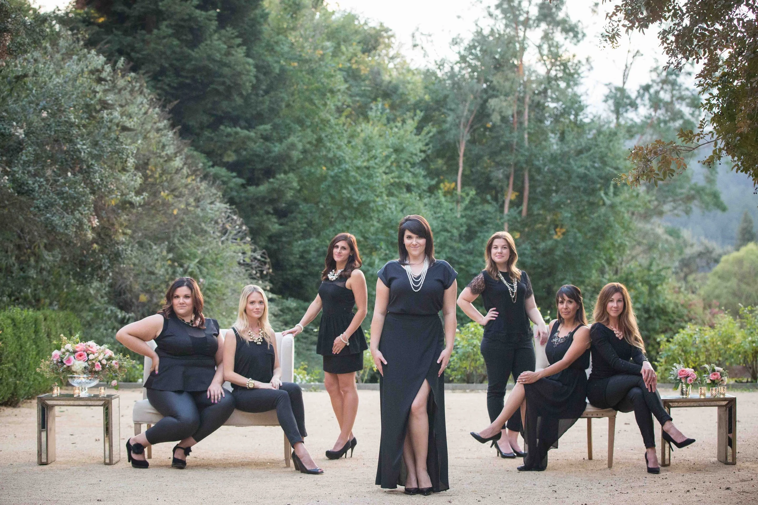 Seven women dressed in black standing and sitting outdoors in a garden or park setting, with trees and greenery in the background, and flower arrangements on small tables on either side.