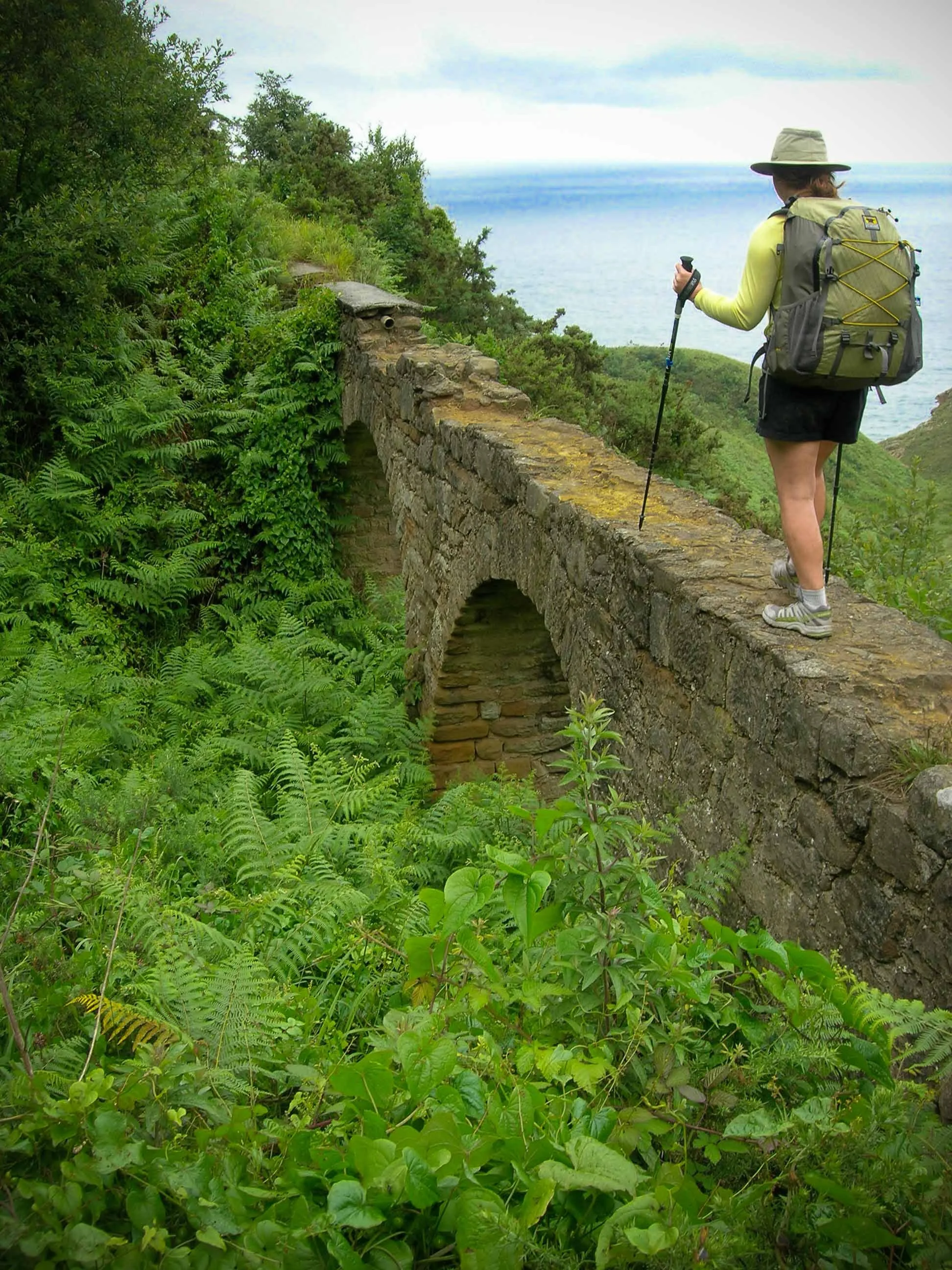 A woman with a backpack and hat hiking along a stone bridge on a lush green hillside overlooking the ocean.