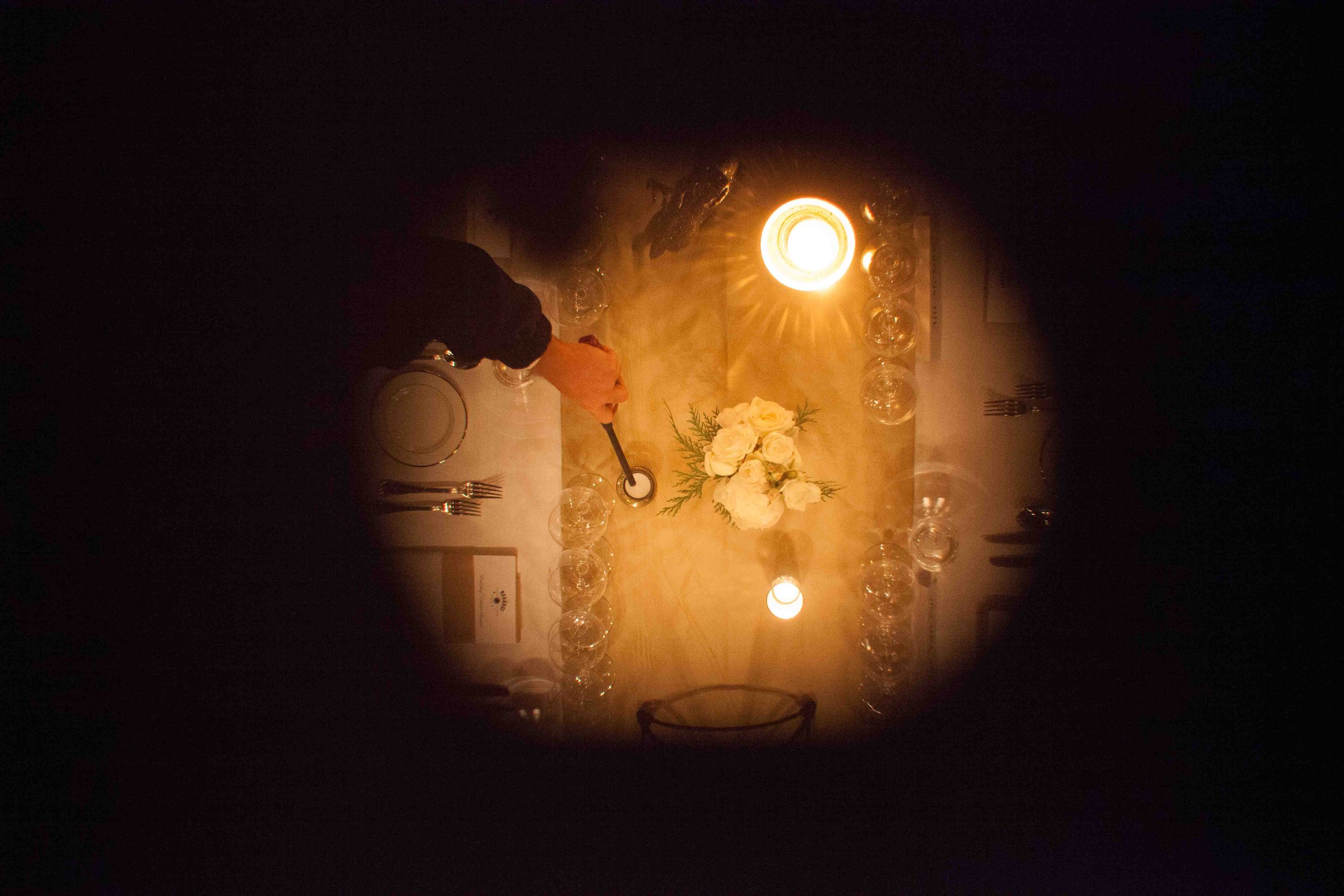 Overhead view of a person lighting a candle at a formal dinner table with white floral centerpiece and glassware, illuminated by warm overhead light.