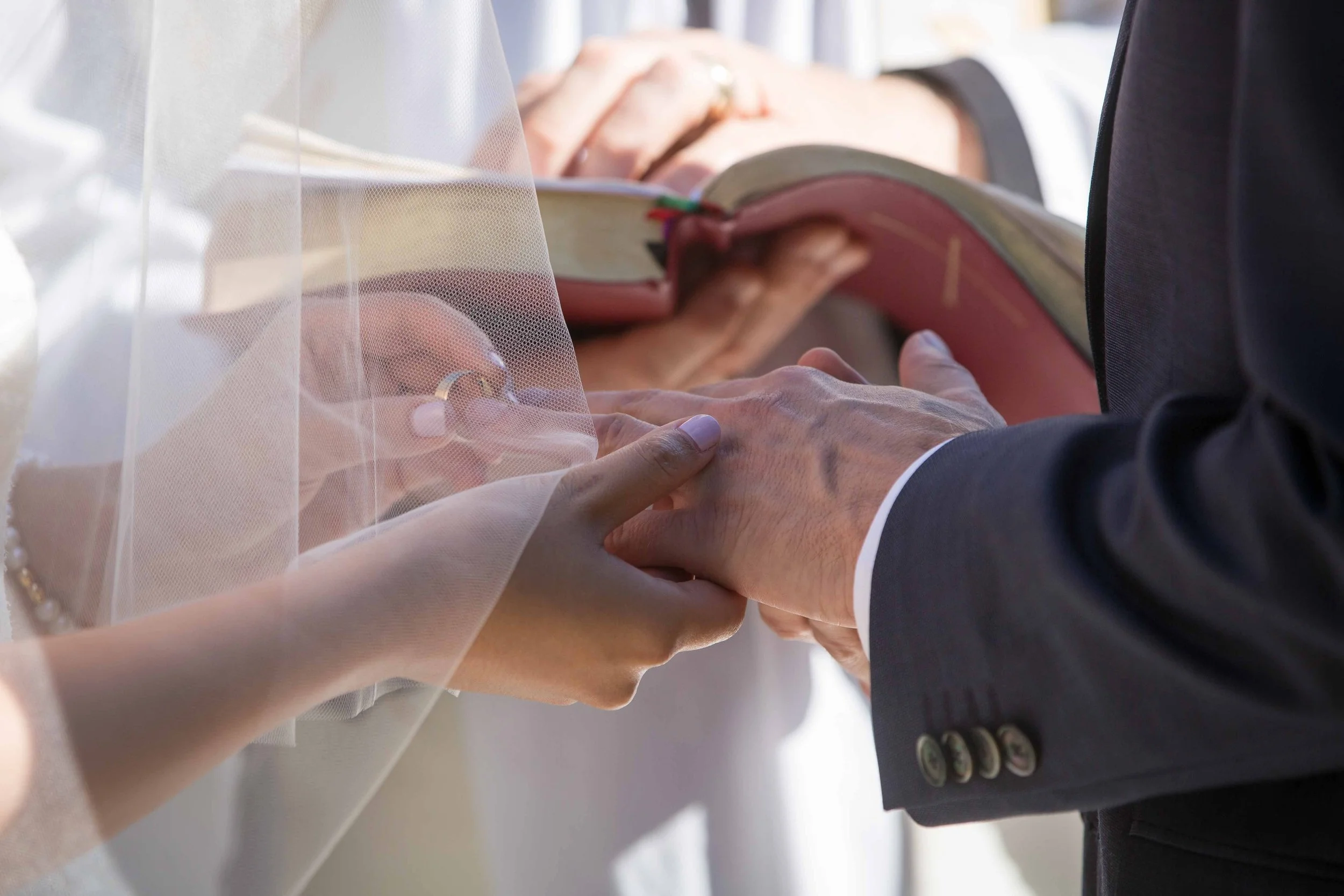 Close-up of a wedding ceremony with a bride and groom exchanging rings, with the bride holding a Bible and wearing a veil.
