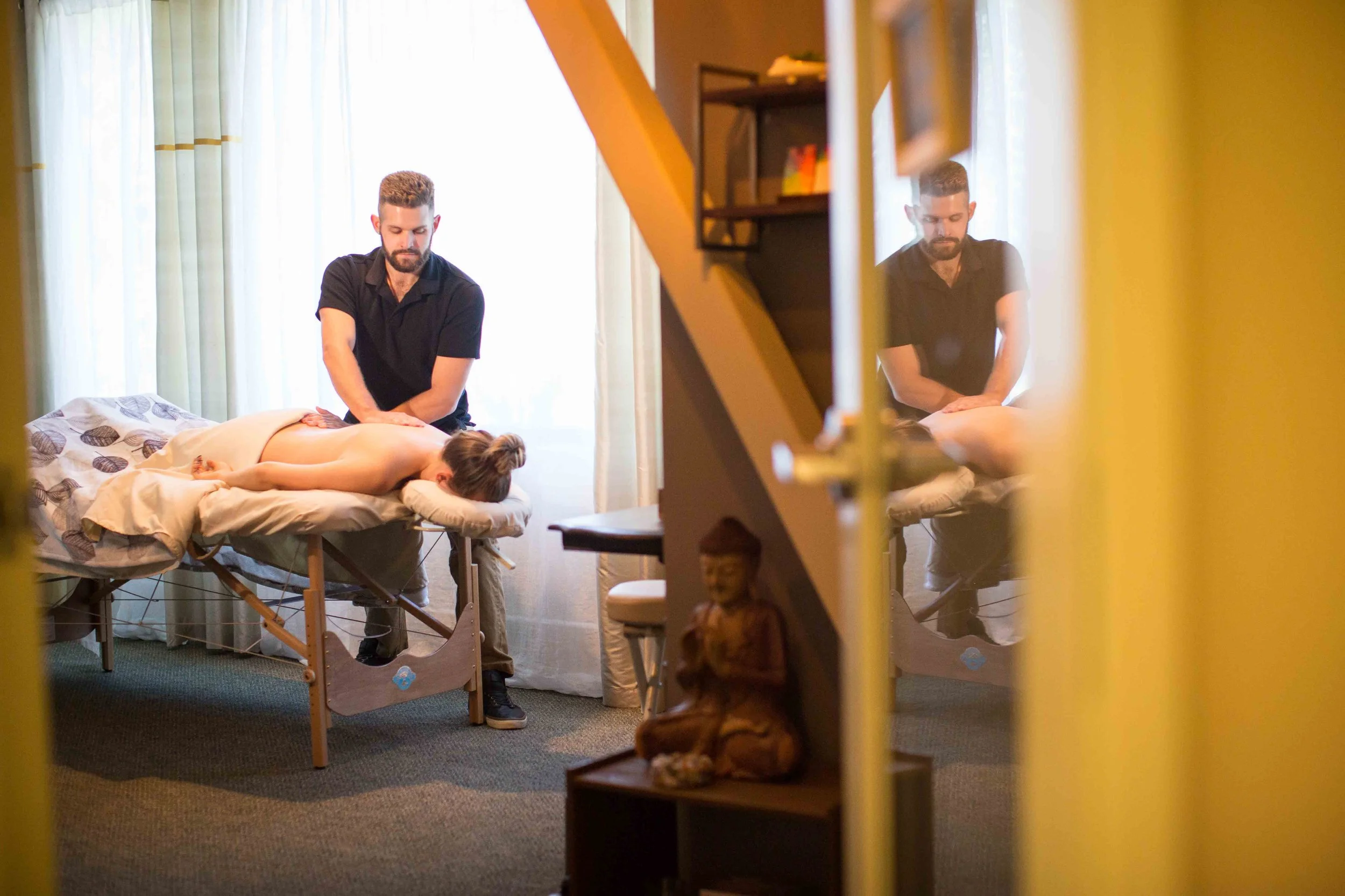 A person receiving a massage while lying face down on a massage table, with a massage therapist standing beside them, in a room with natural light and curtains. The image is viewed through an open door with a reflection of the scene in a mirror.