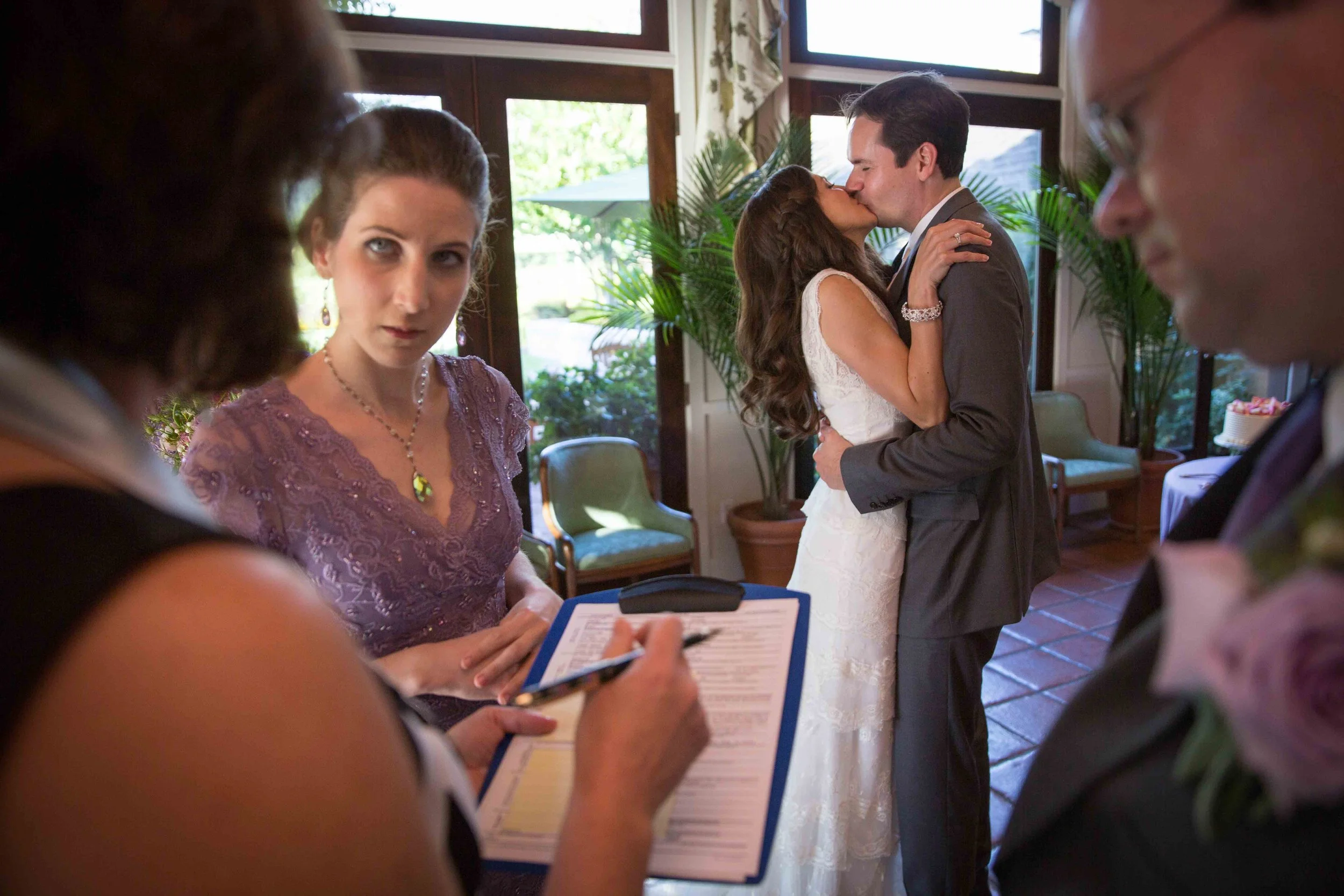 A couple is kissing at their wedding ceremony while two people stand nearby, one holding a clipboard and pen, in a room with large windows and green plants.