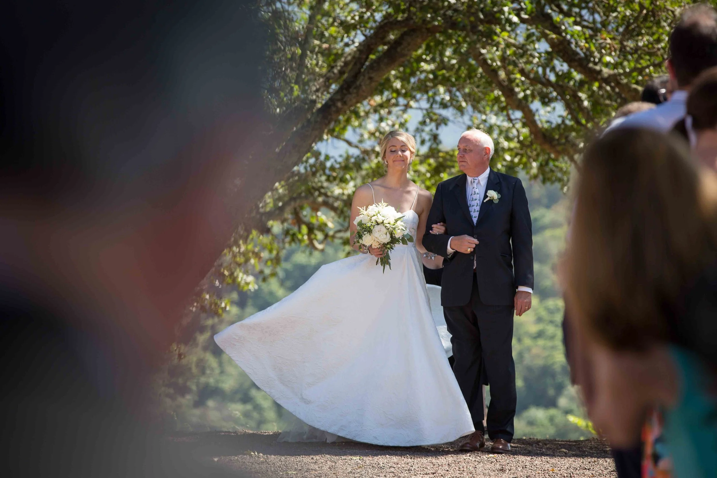 A bride in a white wedding gown holding a bouquet walks down the aisle escorted by an older man in a dark suit, outdoors under a tree on a sunny day, with guests visible on the sides.