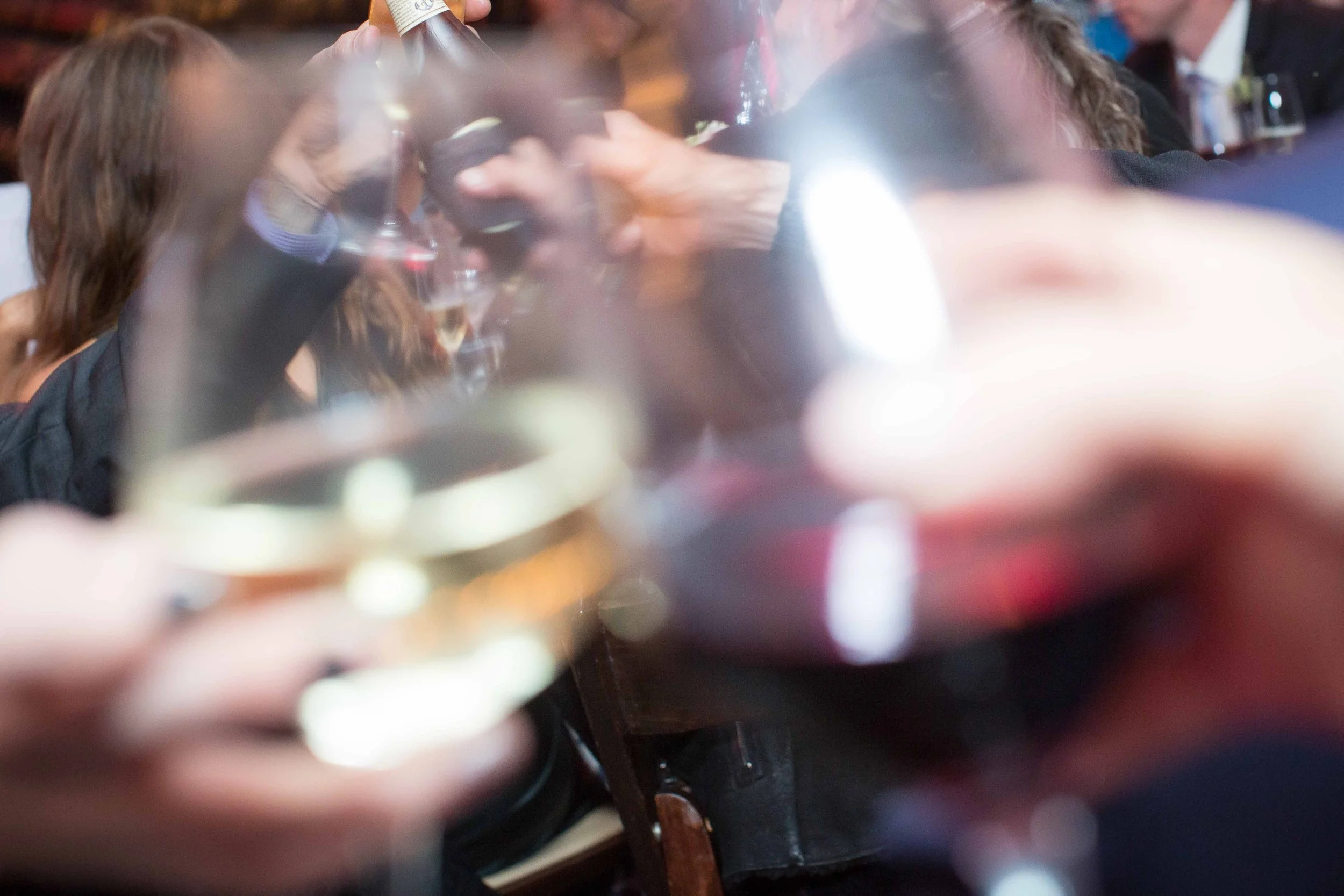 A blurry photo of people at a social gathering, some holding glasses of wine or champagne, seen through a glass or reflective surface.