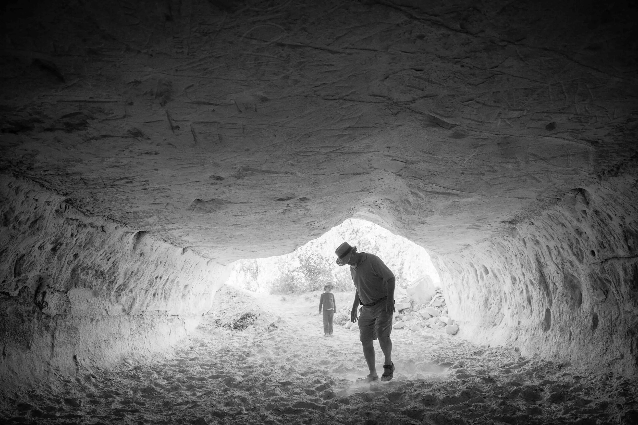 A man and a child walking through a cave with light shining in from the entrance, black and white photo.