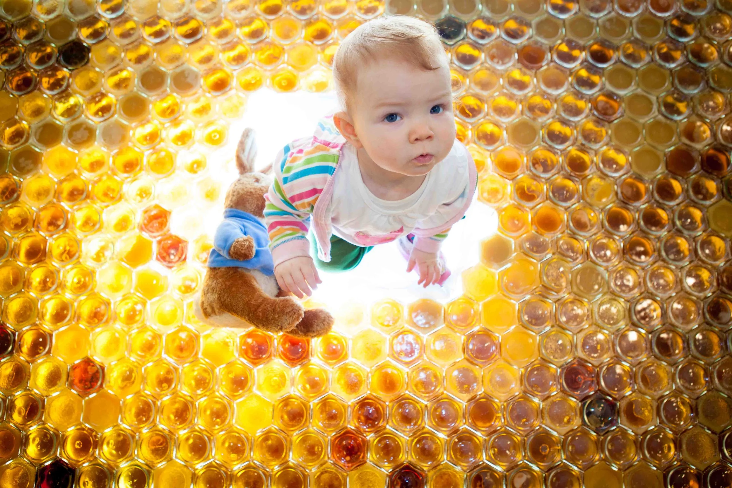 Young child with blonde hair, wearing a white shirt with colorful stripes and green pants, crawling on a honeycomb-patterned surface filled with honey, with a plush teddy bear next to him.