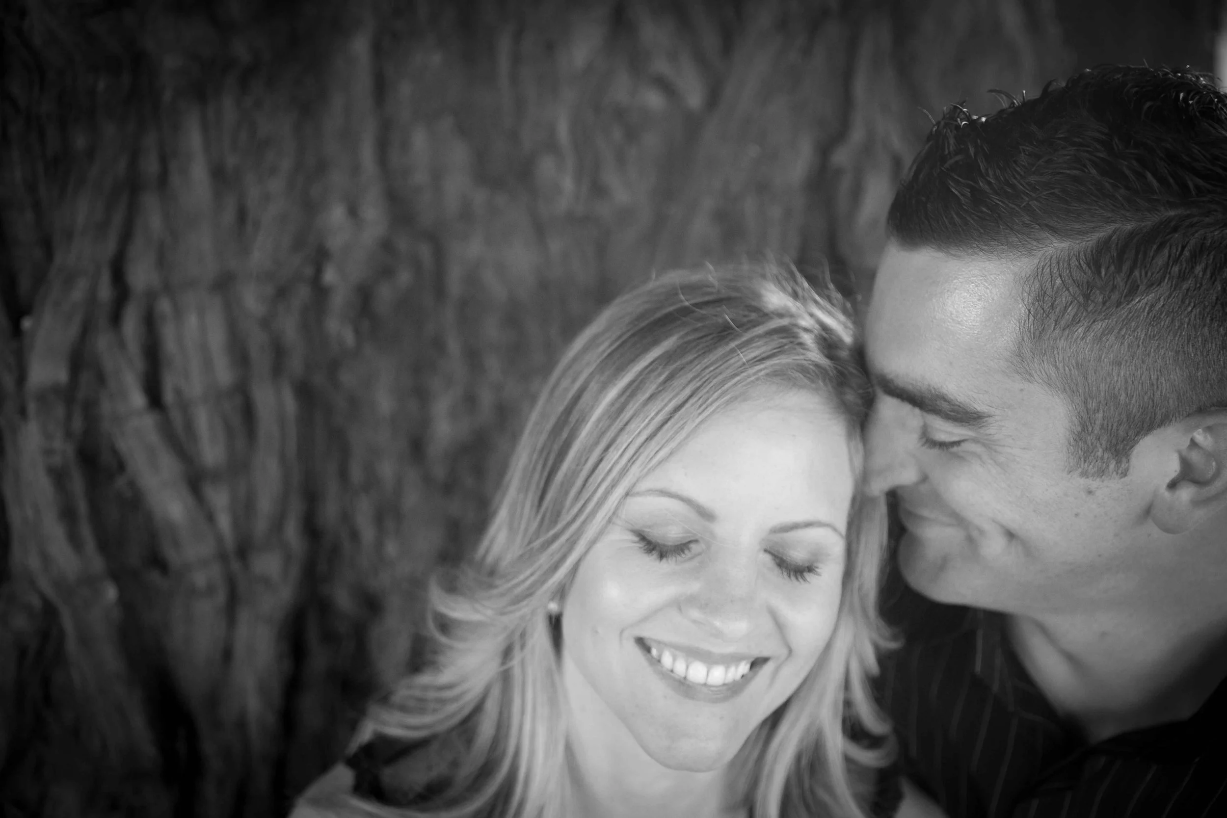 A black and white photo of a smiling woman with closed eyes and a man close to her, both with their foreheads touching, against a wooden background.