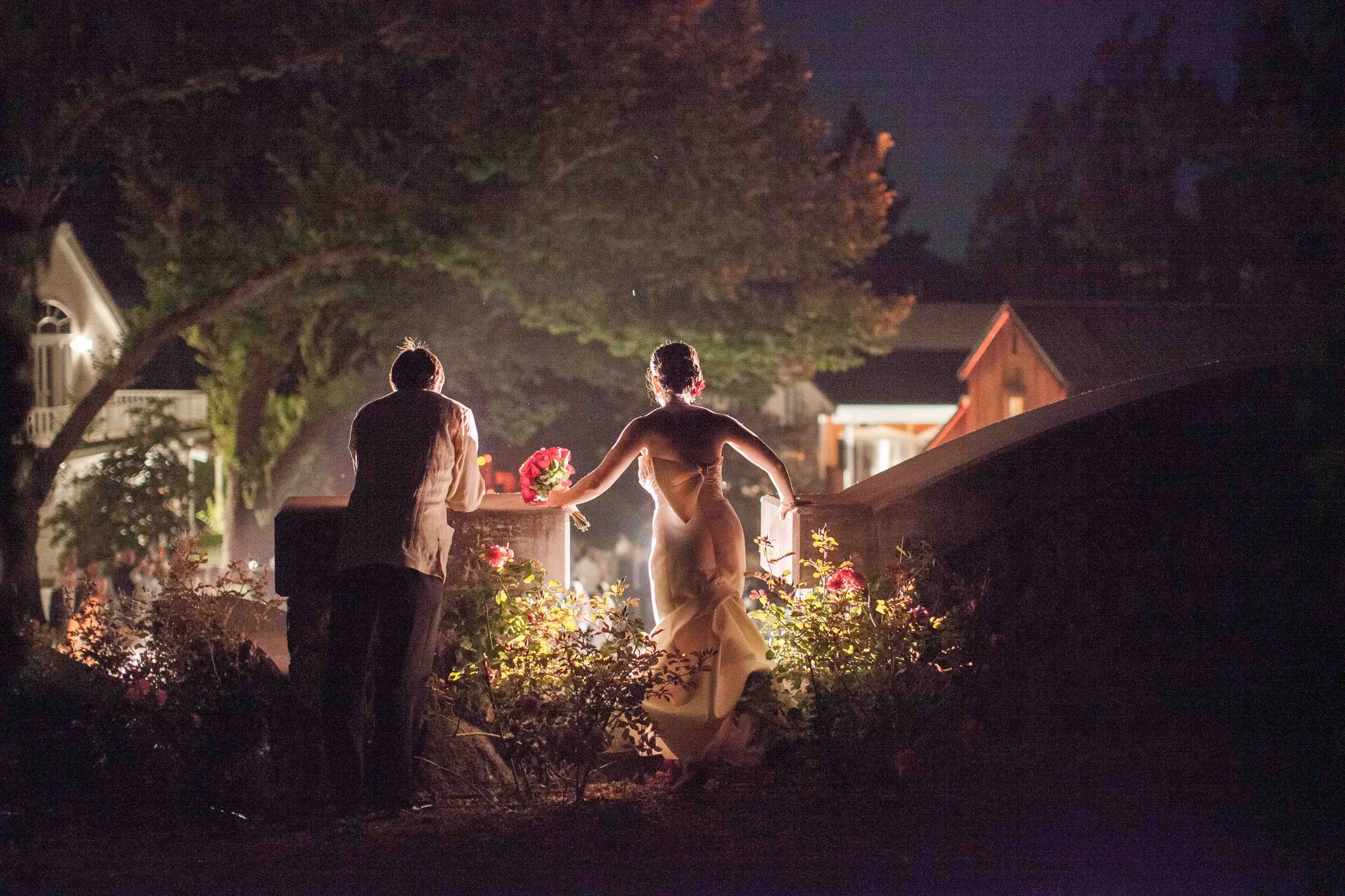 A couple stands on a bridge at night, with the man on the left and the woman on the right, holding a bouquet of flowers, illuminated by warm light in a suburban neighborhood.