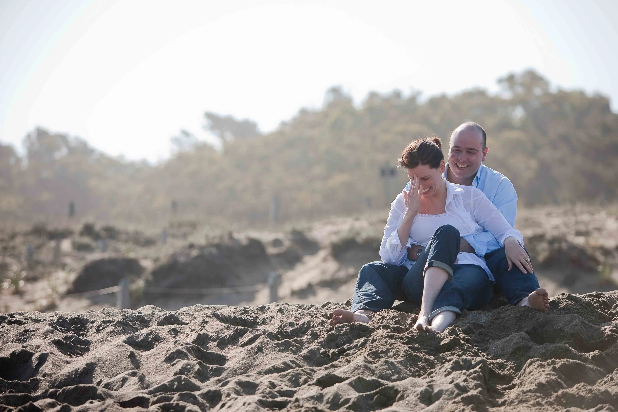 A smiling couple sitting on sandy ground outdoors, with trees and sky in the background, sharing a joyful moment.