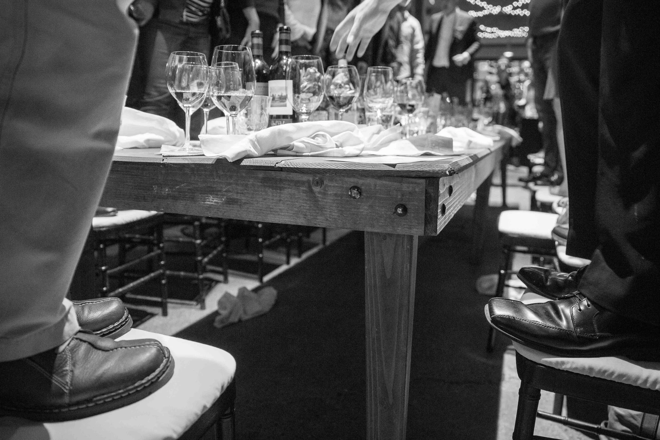 A long wooden table set with multiple glasses, bottles, and napkins, surrounded by chairs with people standing or sitting around in a lively indoor gathering or party.
