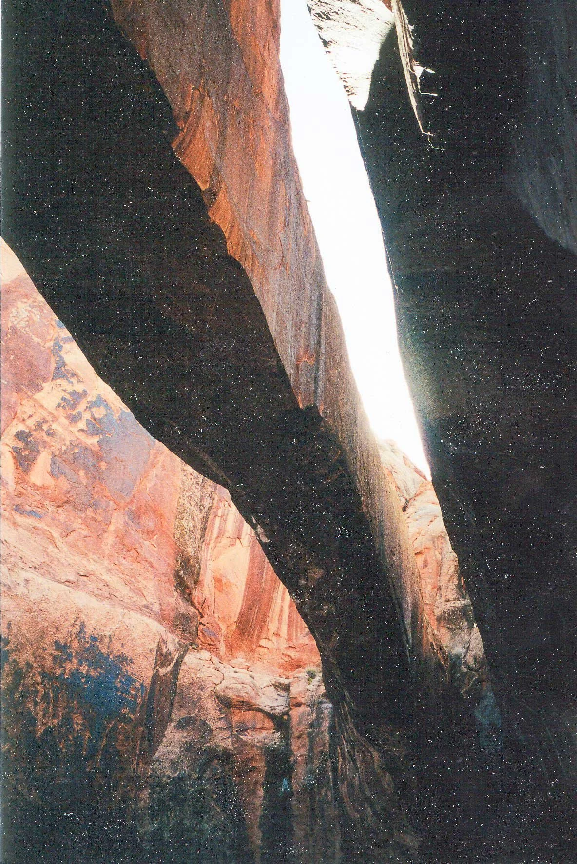 A narrow, winding canyon with steep, colorful rock walls and a small waterfall or stream flowing at the top of the canyon.