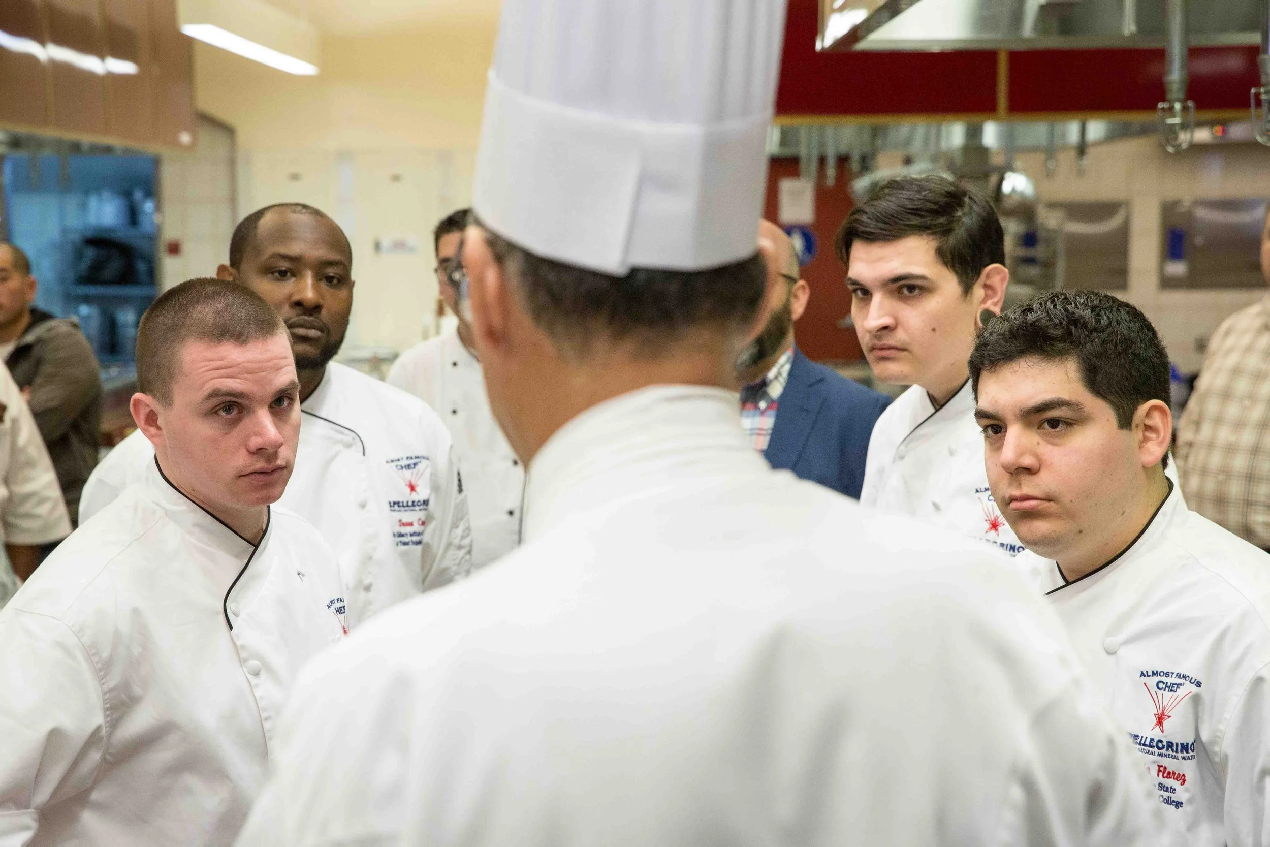 A group of young male culinary students listening to an instructor in a professional kitchen, all wearing white chef uniforms and hats.