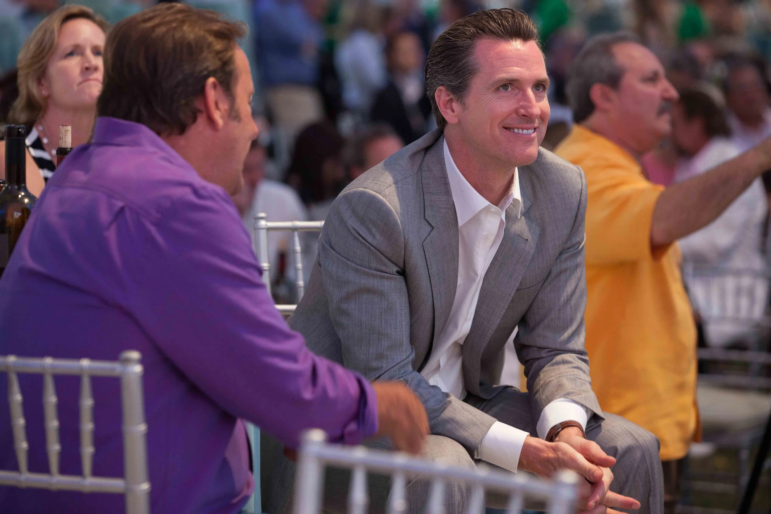 Three people sitting in chairs at an outdoor event, with a man in a gray suit smiling and looking to his right, a woman in an orange shirt waving, and a person in purple in profile.