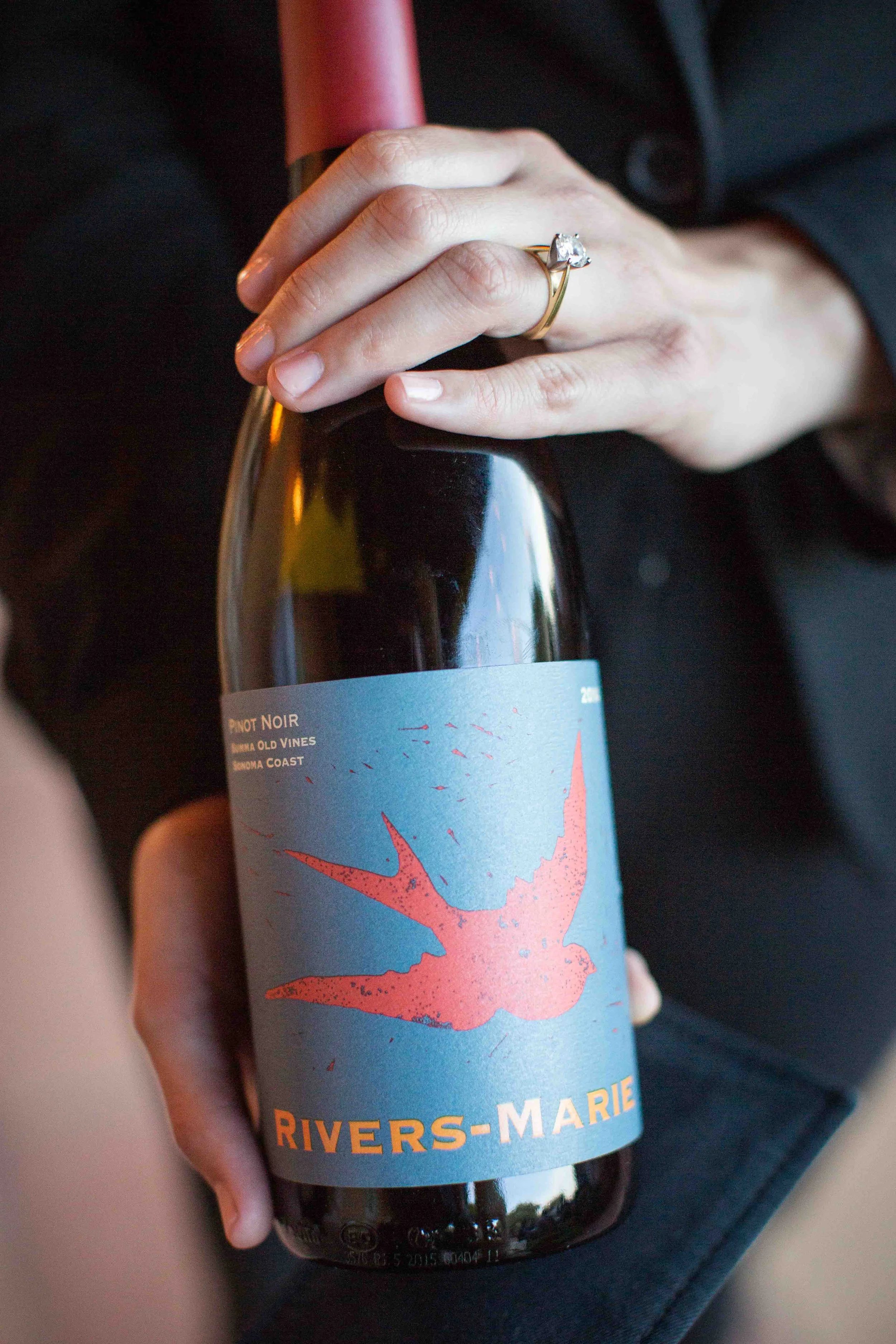 Person holding a bottle of Rivers-Marie Pinot Noir wine with a pink candle near the neck, and a gold ring with a large gemstone on their finger.