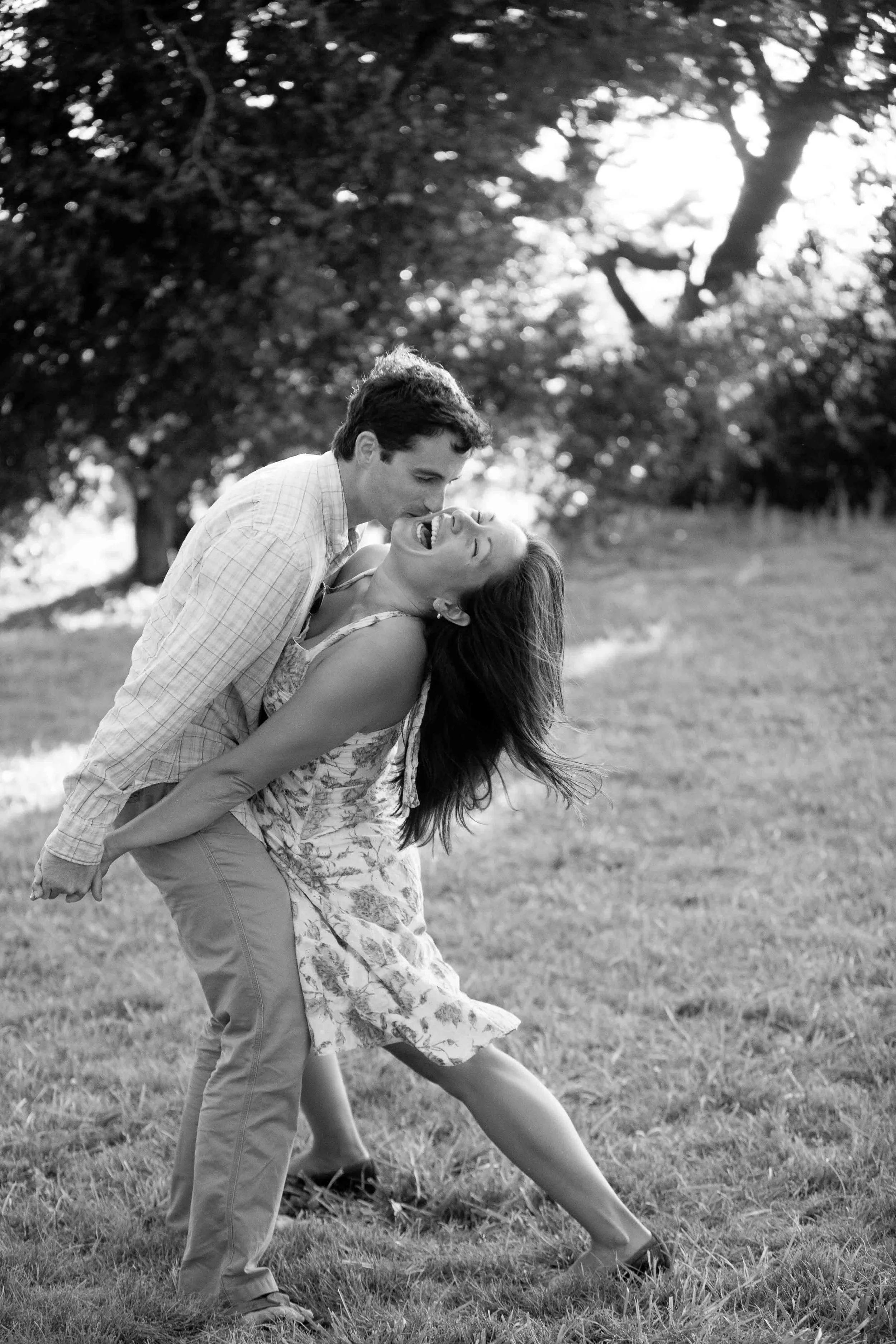 A man and woman outdoors, the man is giving the woman a piggyback ride, both laughing and enjoying themselves, surrounded by trees in a park.