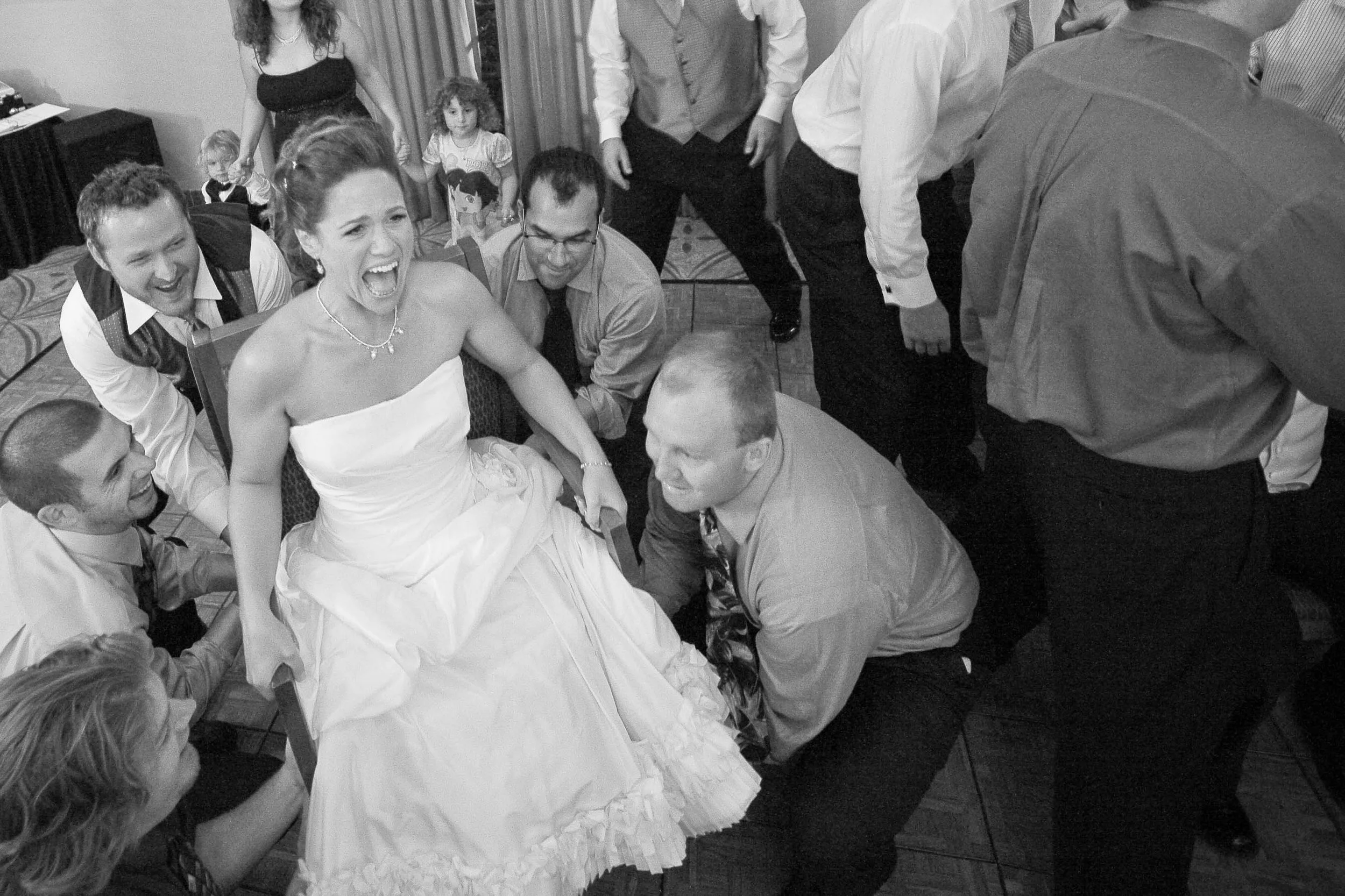 A woman in a wedding dress sitting on a chair, laughing loudly, surrounded by men and women in formal attire at a wedding reception, with a joyful atmosphere.