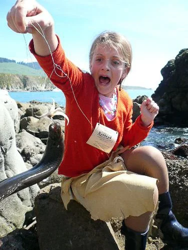 Child sitting on rocks near the water, holding a snake with a surprised expression, wearing an orange sweater and beige shorts.