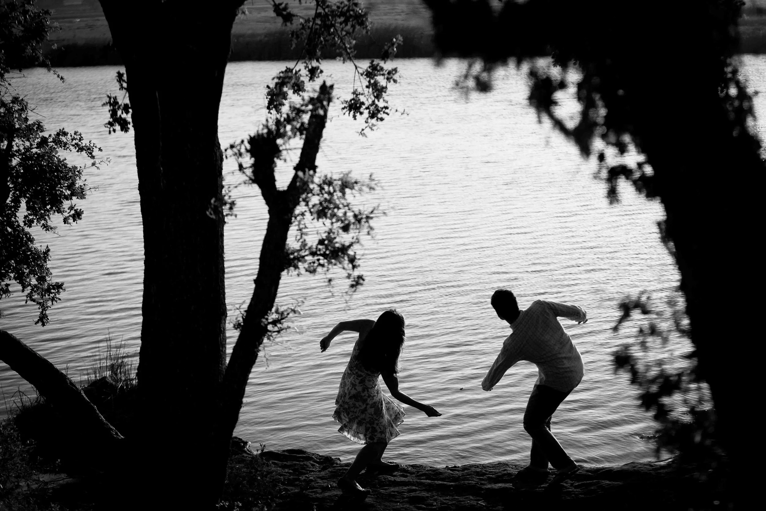 Two children, a girl and a boy, playing by a lake at sunset, surrounded by trees.