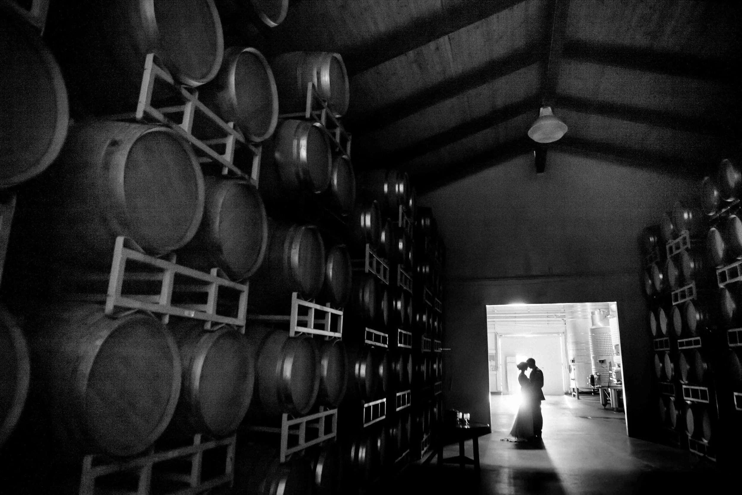 Silhouettes of two people standing and talking inside a wine cellar with stacked barrels on the walls.